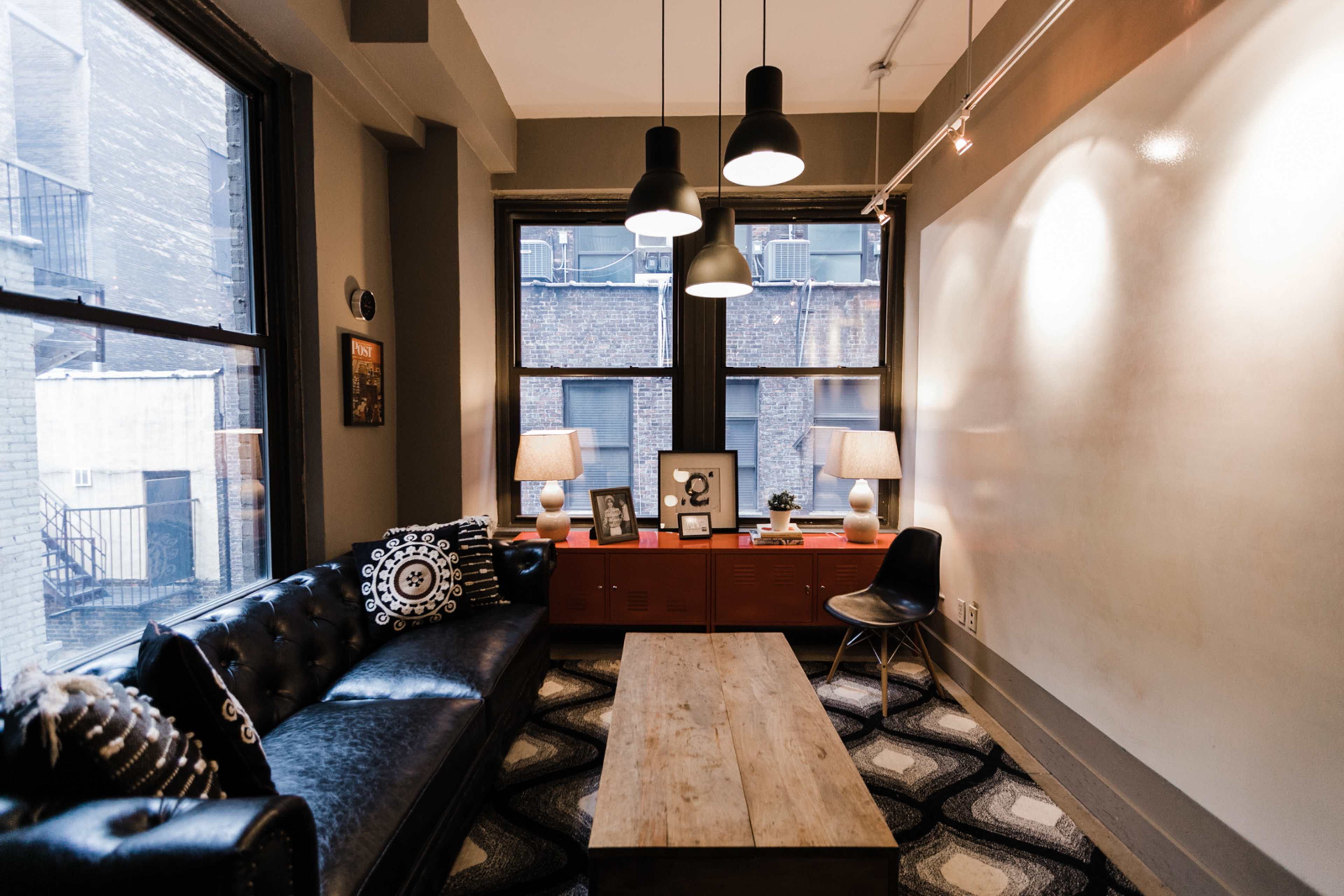 The image shows a stylish interior of a cozy living room featuring a black leather sofa, wooden coffee table, and large windows with a view of an adjacent building.