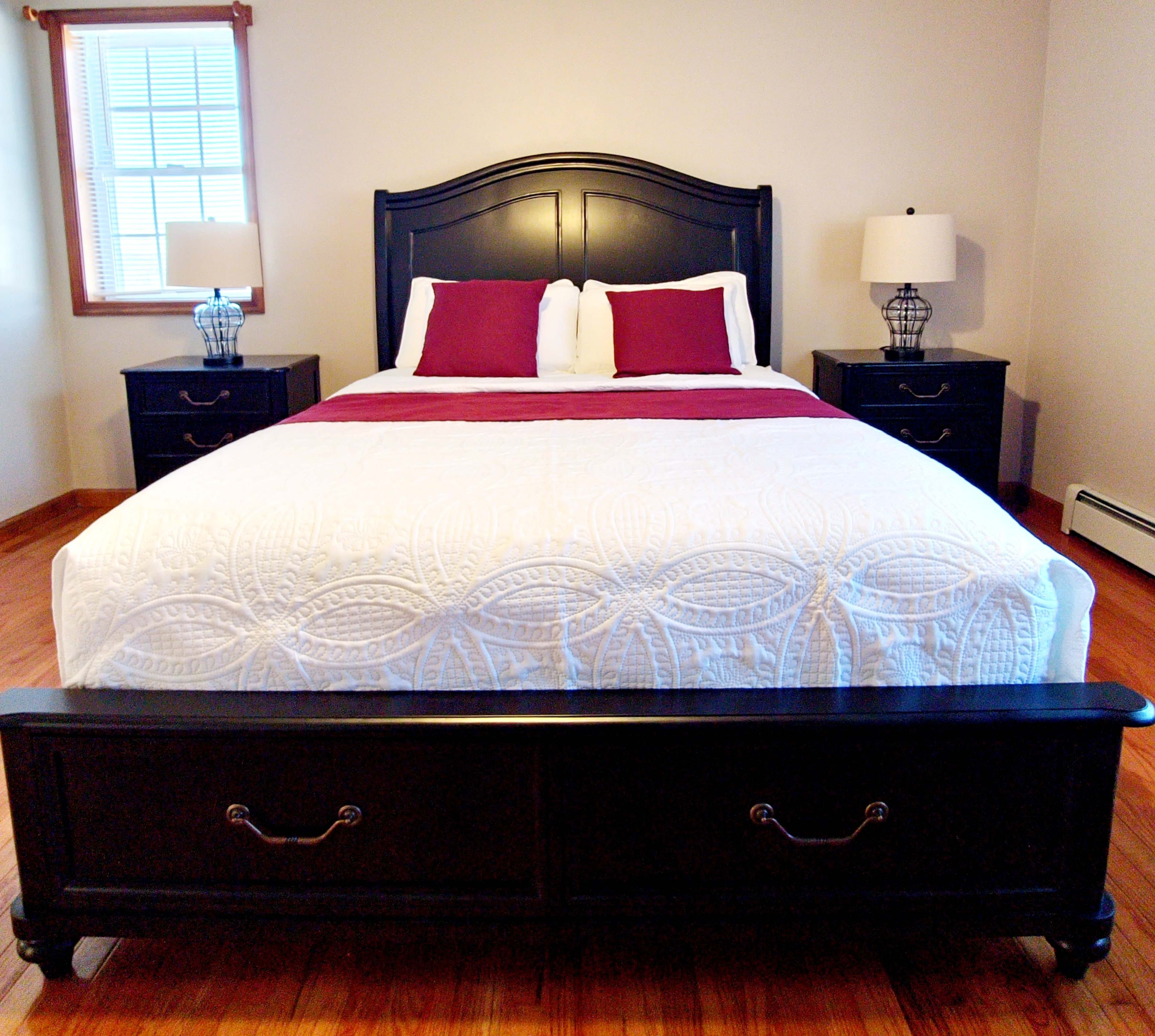 A bedroom featuring a large black bed with a white quilted bedspread and burgundy accents, flanked by two nightstands with lamps.