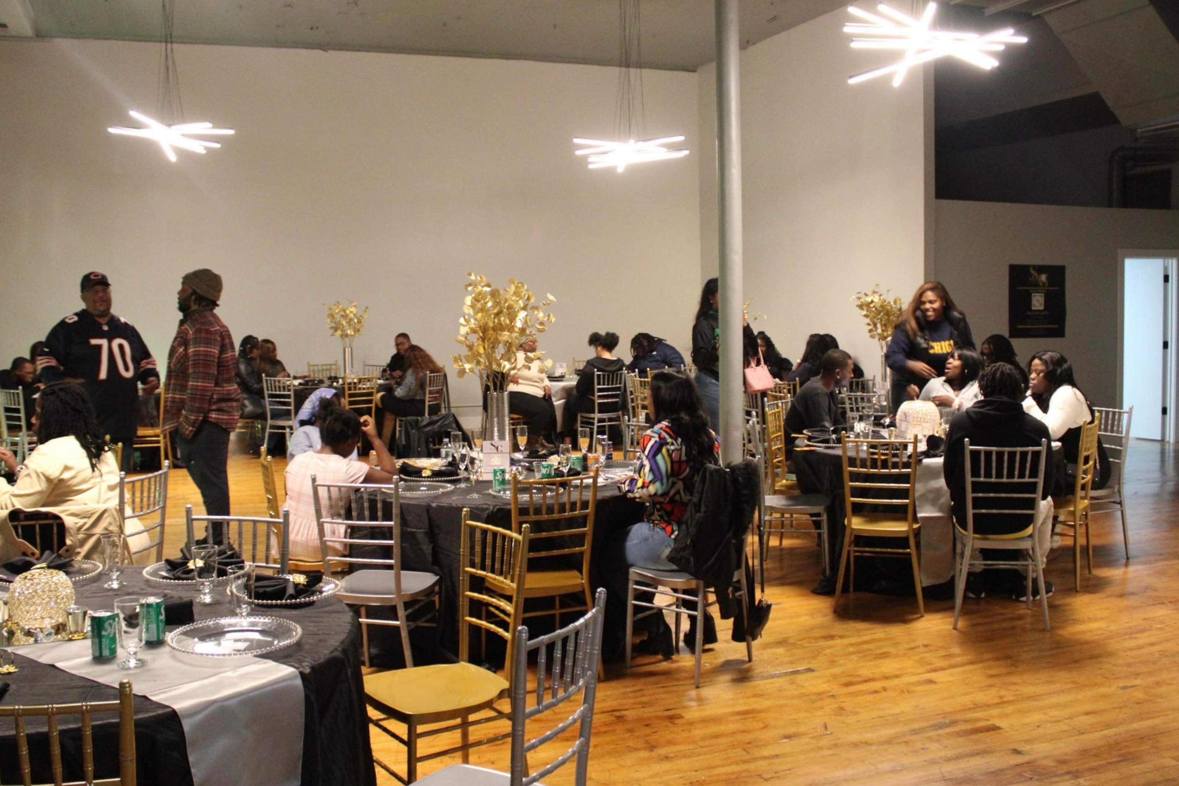 A large room features several tables set for a gathering, with guests seated and others mingling, surrounded by decorative centerpieces.