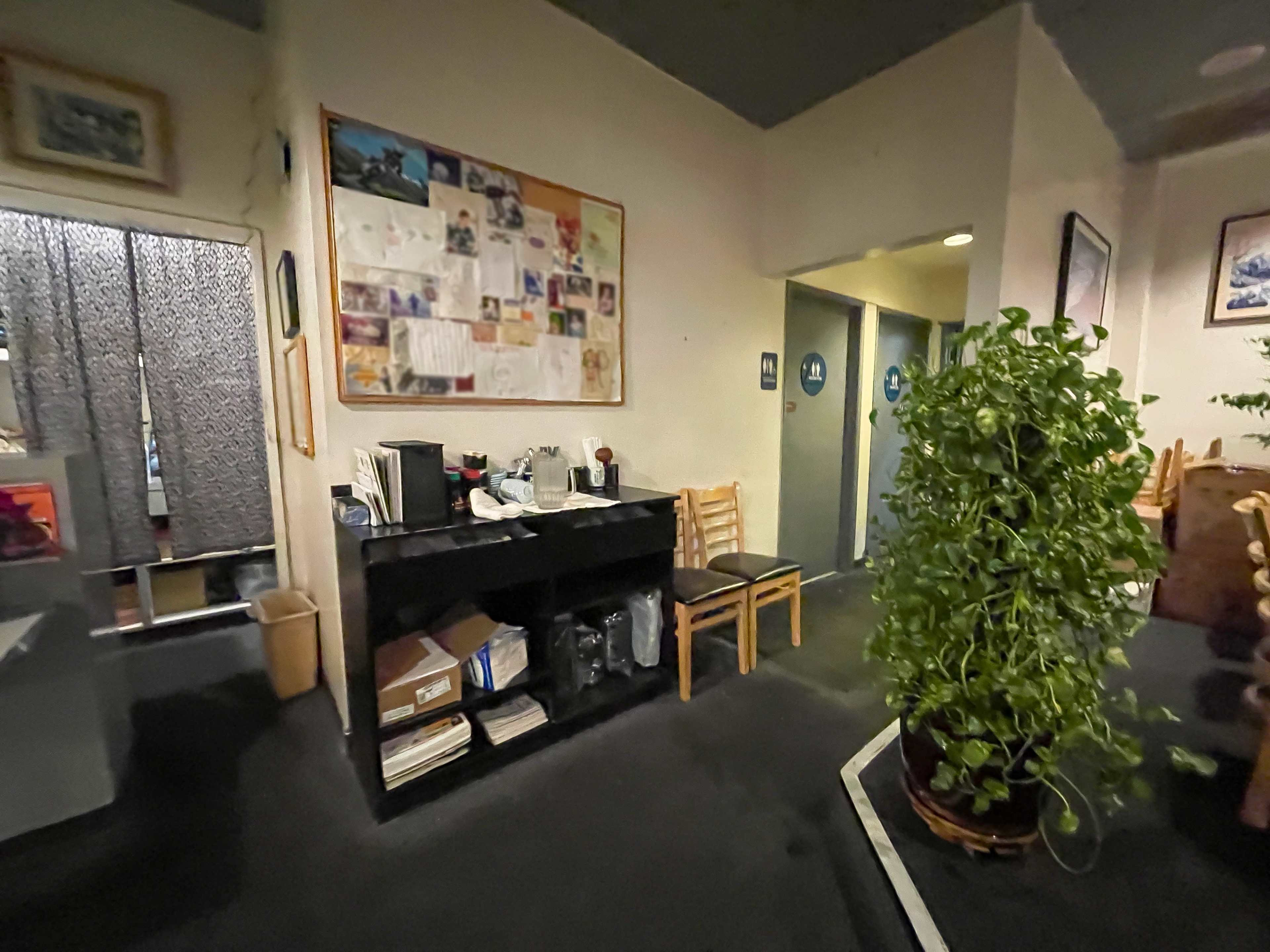 The image shows a small, cluttered reception area with a black countertop, chairs, a potted plant, and two restroom doors at the far end.