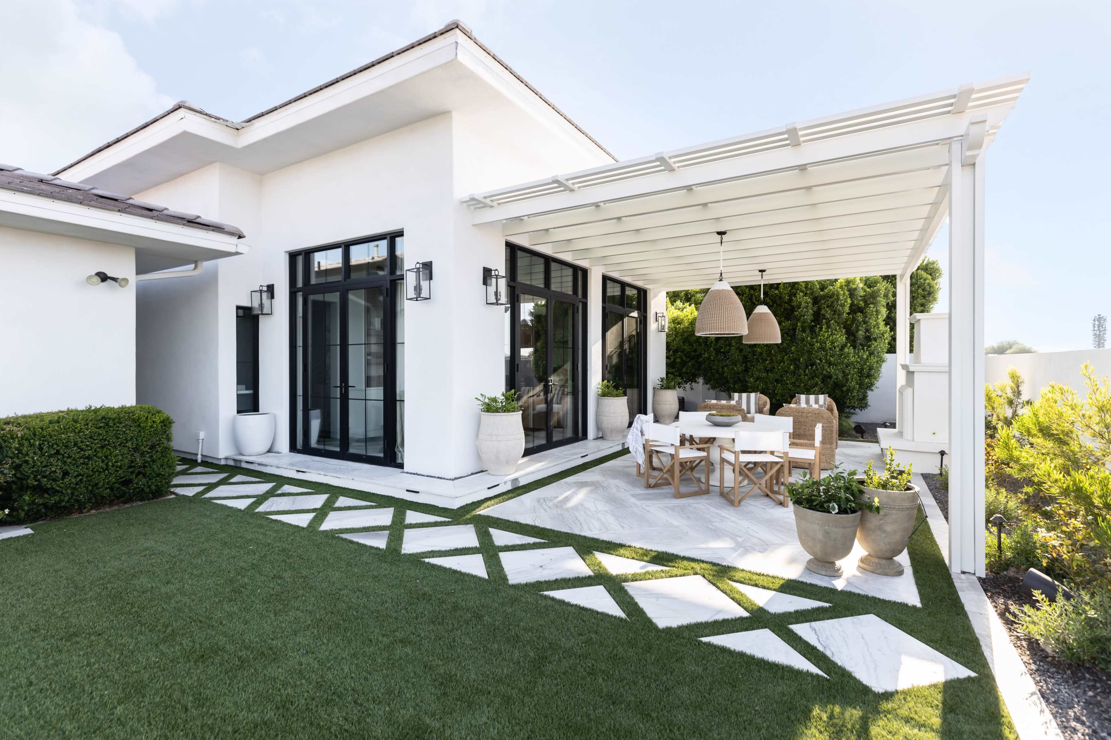 A modern patio features white furniture and pendant lights beneath a covered area, surrounded by neatly arranged grass and marble pathways.