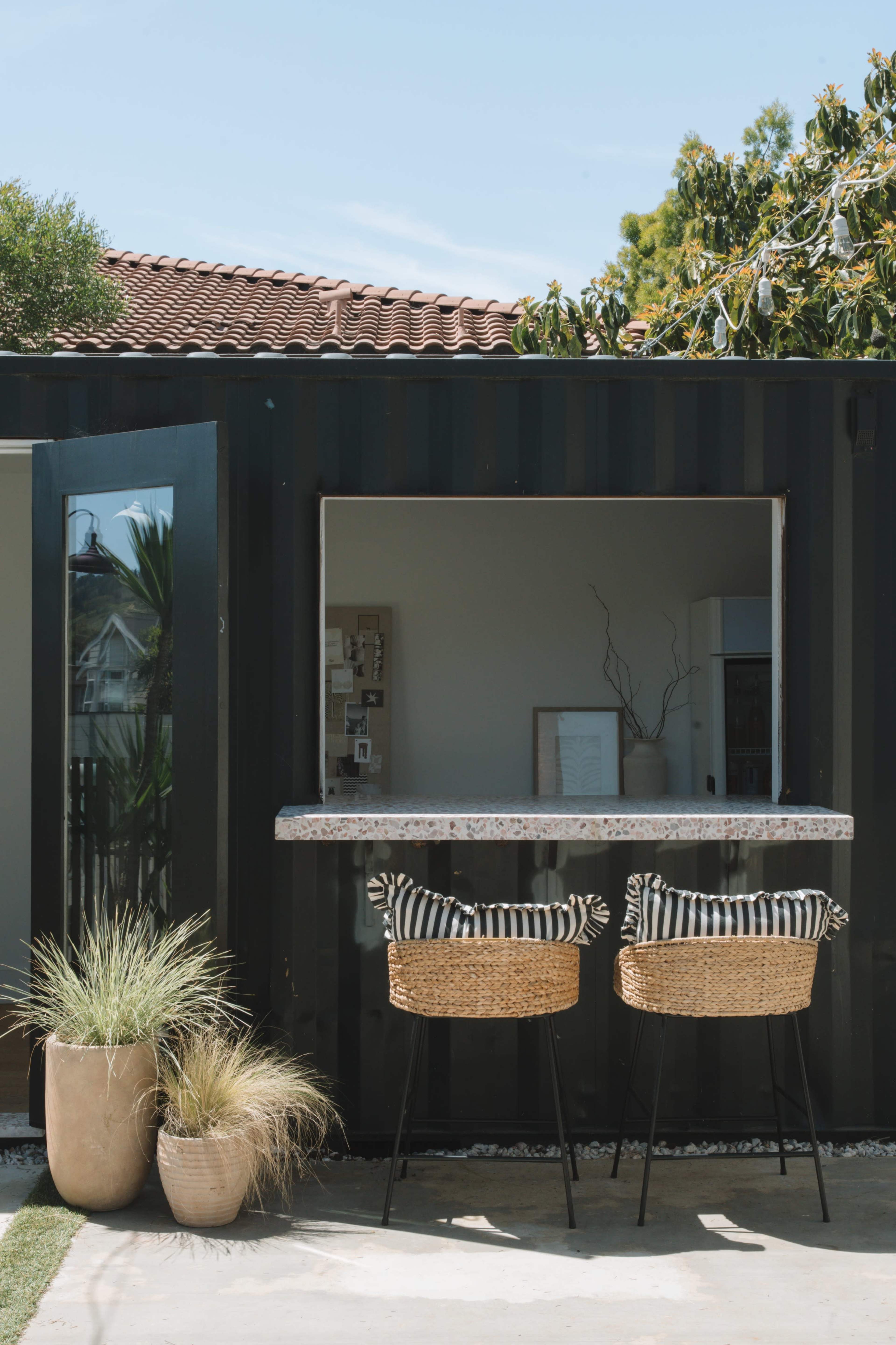 A small outdoor bar area features two woven chairs with cushions beside a large window in a shipping container structure, complemented by potted plants.