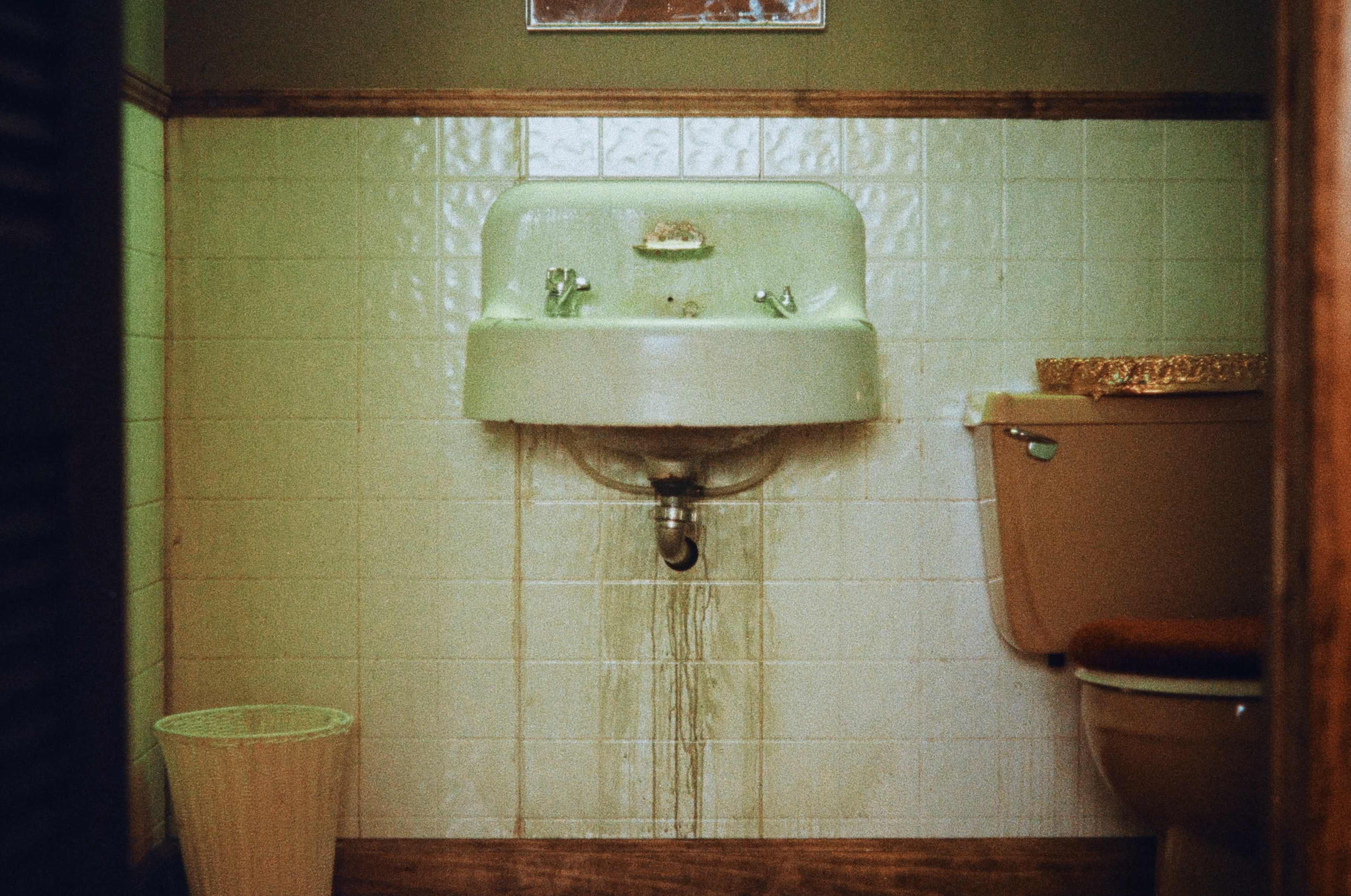 The image shows a bathroom with a green sink and a toilet, featuring leaking water from the sink onto the tiled floor.