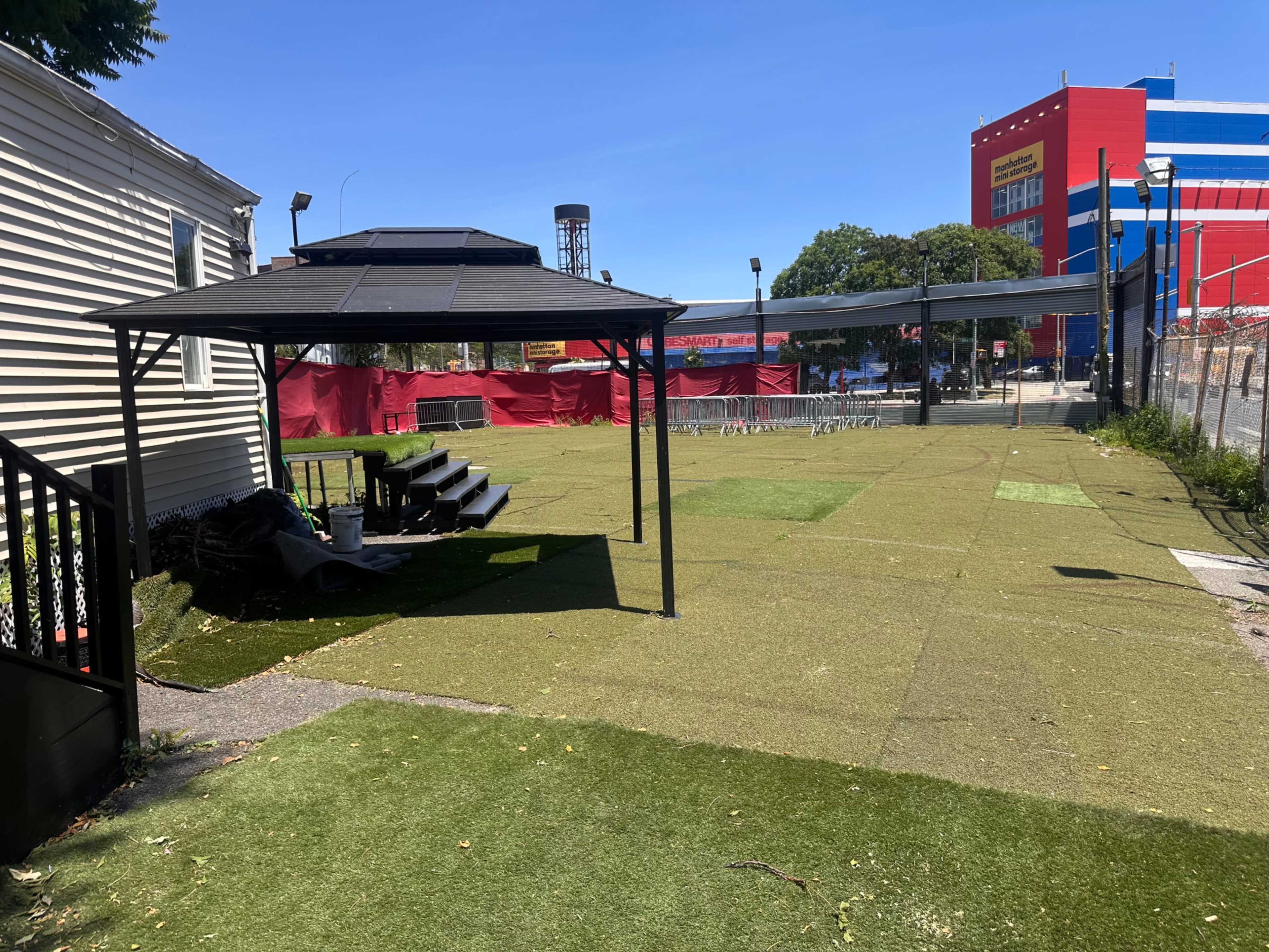 The image shows a fenced outdoor area with artificial grass, featuring a shaded seating area and visible structures in the background.