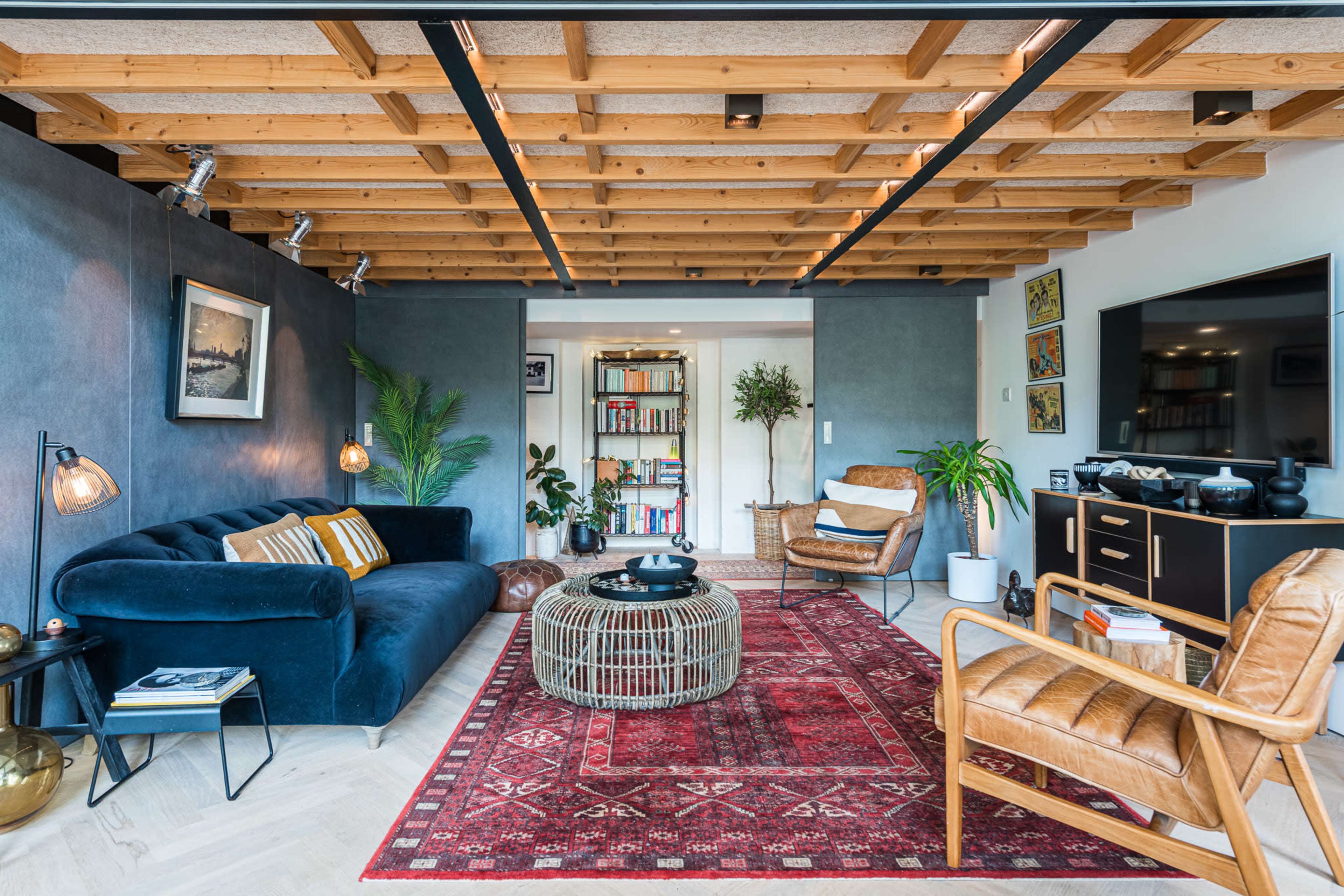 The image shows a modern living room featuring a dark blue sofa, a wooden ceiling, a circular coffee table, and a mix of potted plants and artwork.