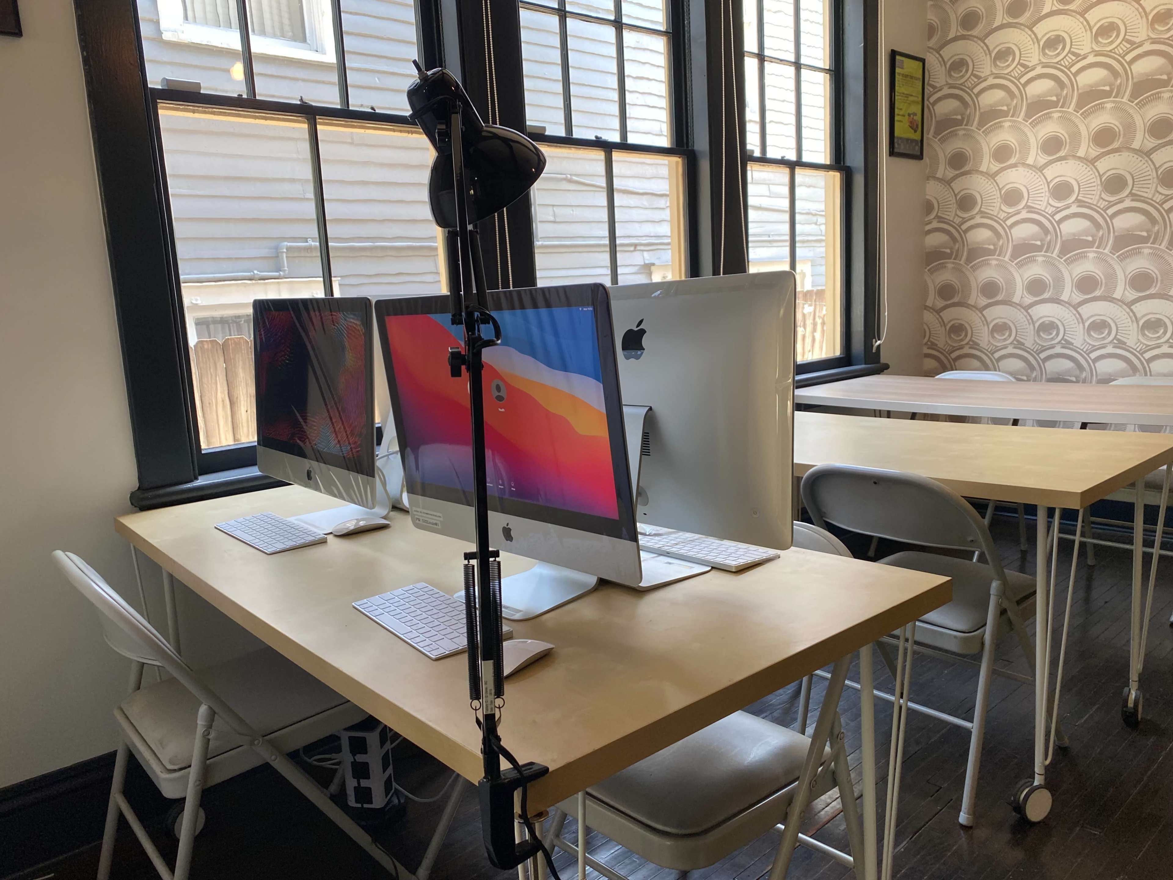 A workspace features two iMac computers on a table, accompanied by folding chairs and a lighting stand.