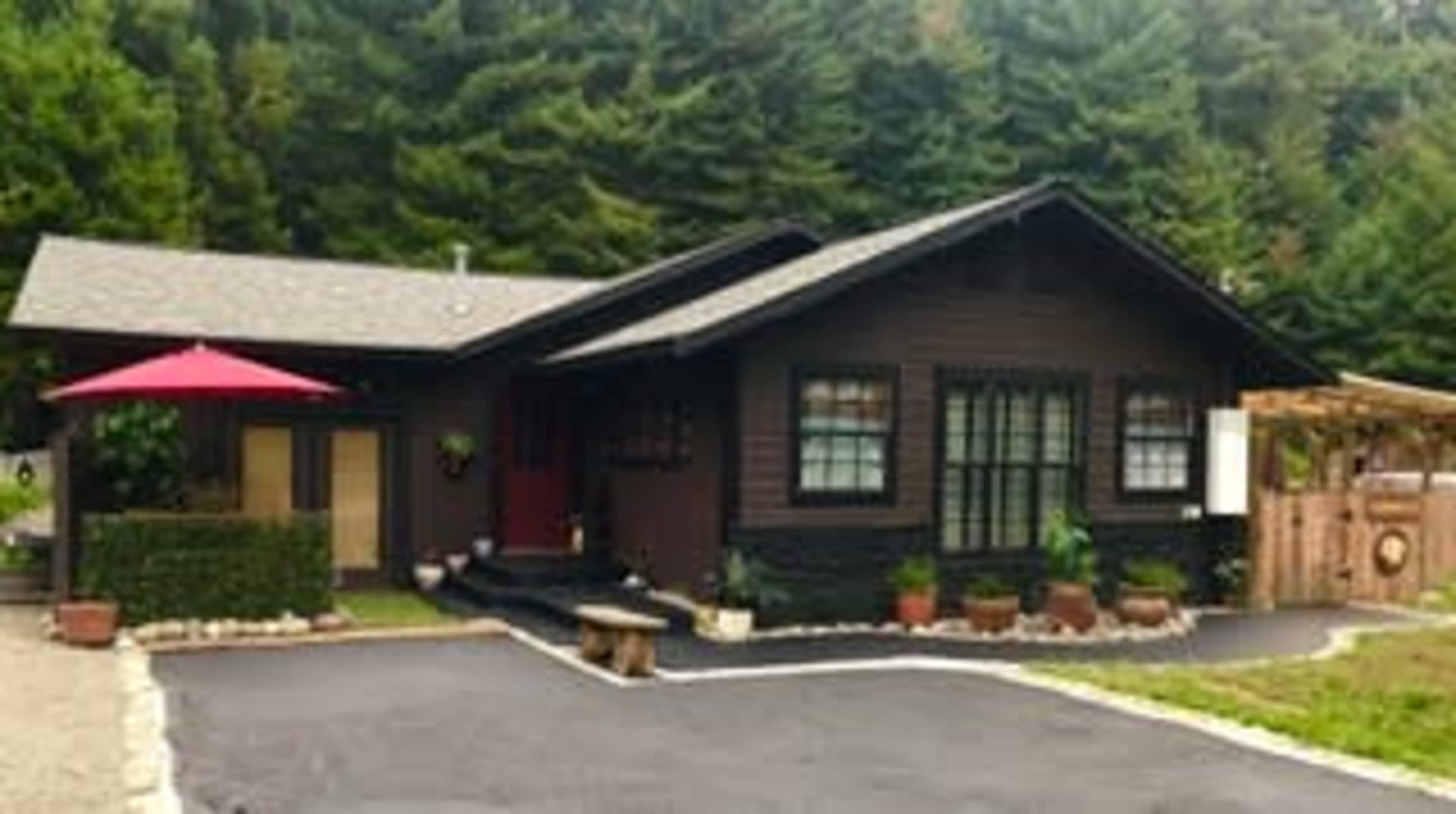 A dark-colored bungalow with a sloped roof is surrounded by greenery and features a red umbrella over a patio area.
