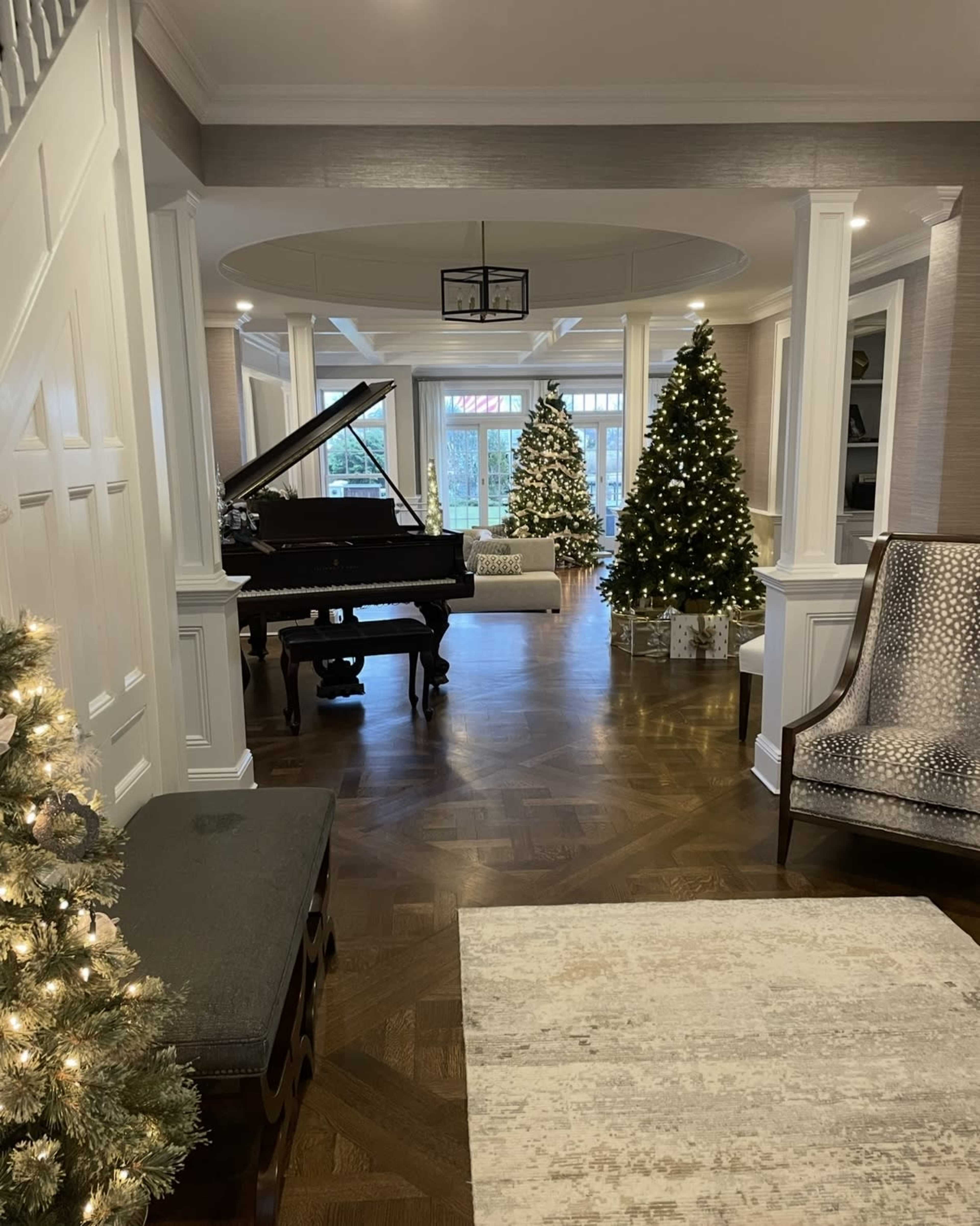 The image shows a cozy entrance hall featuring a grand piano, two Christmas trees adorned with lights, and a patterned area rug on polished wooden flooring.