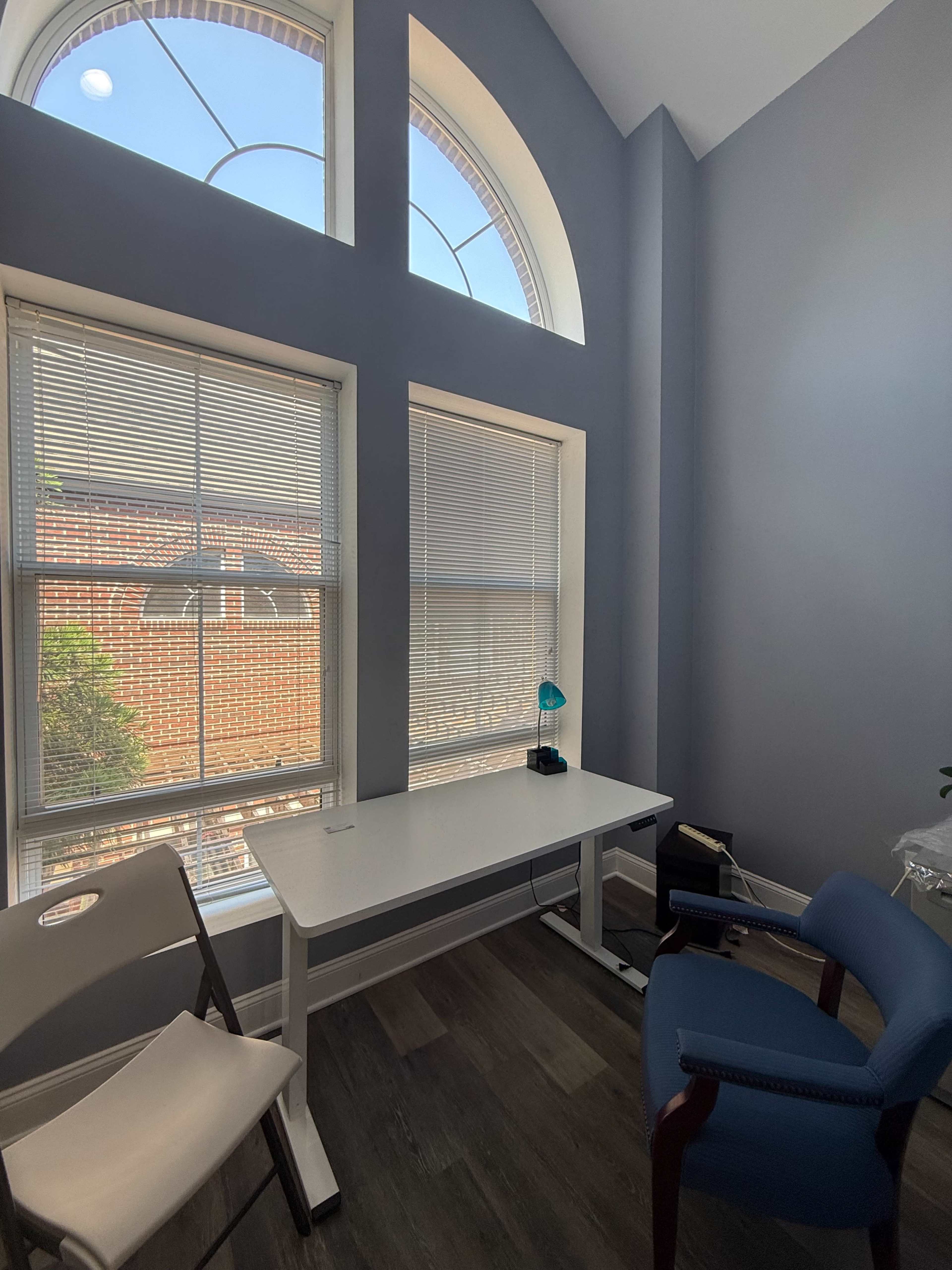 The image shows a small office space with a white desk and a blue chair situated near large windows with vertical blinds.