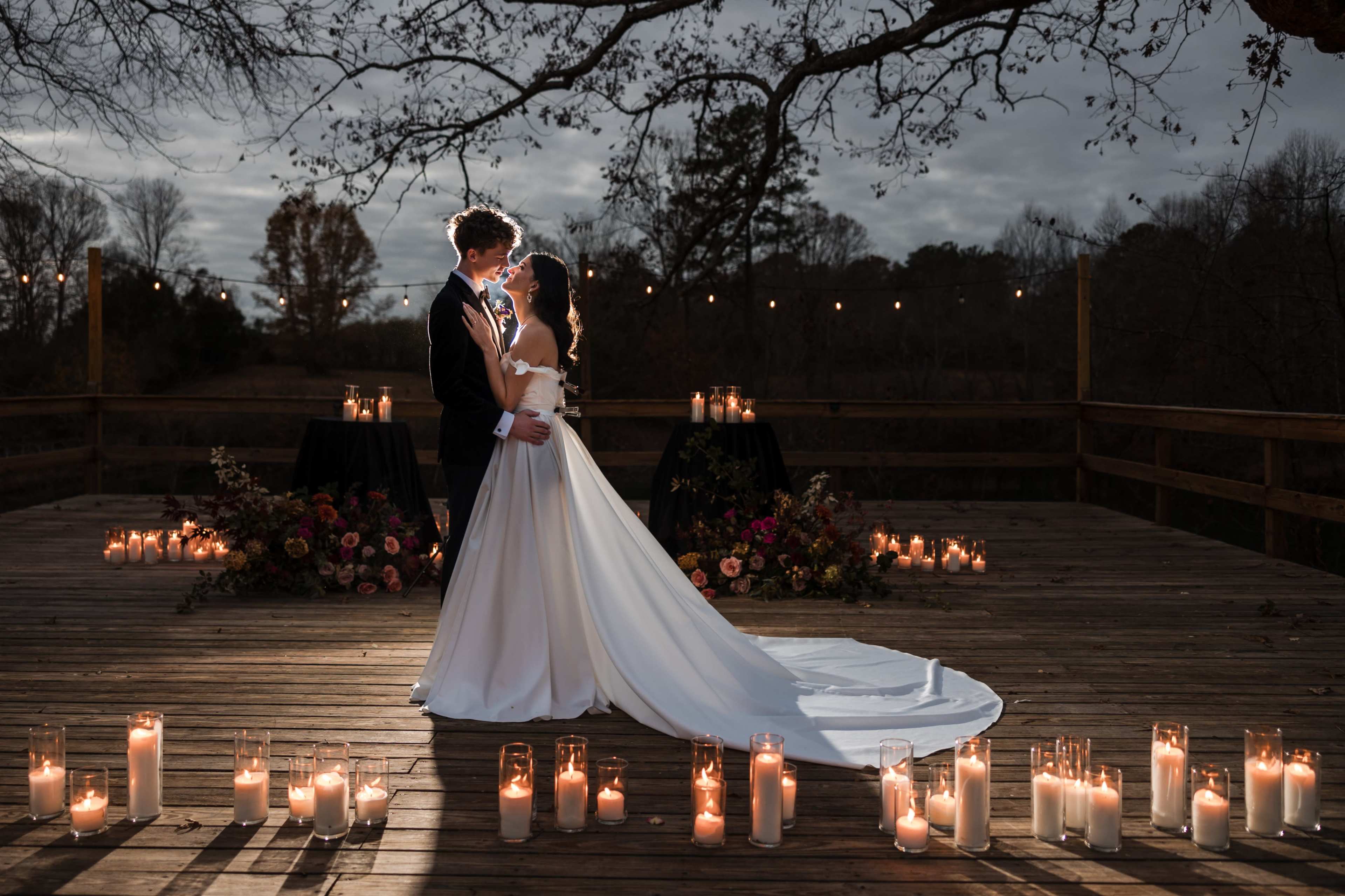A couple in formal wedding attire stands together on a wooden deck surrounded by candles and floral arrangements.