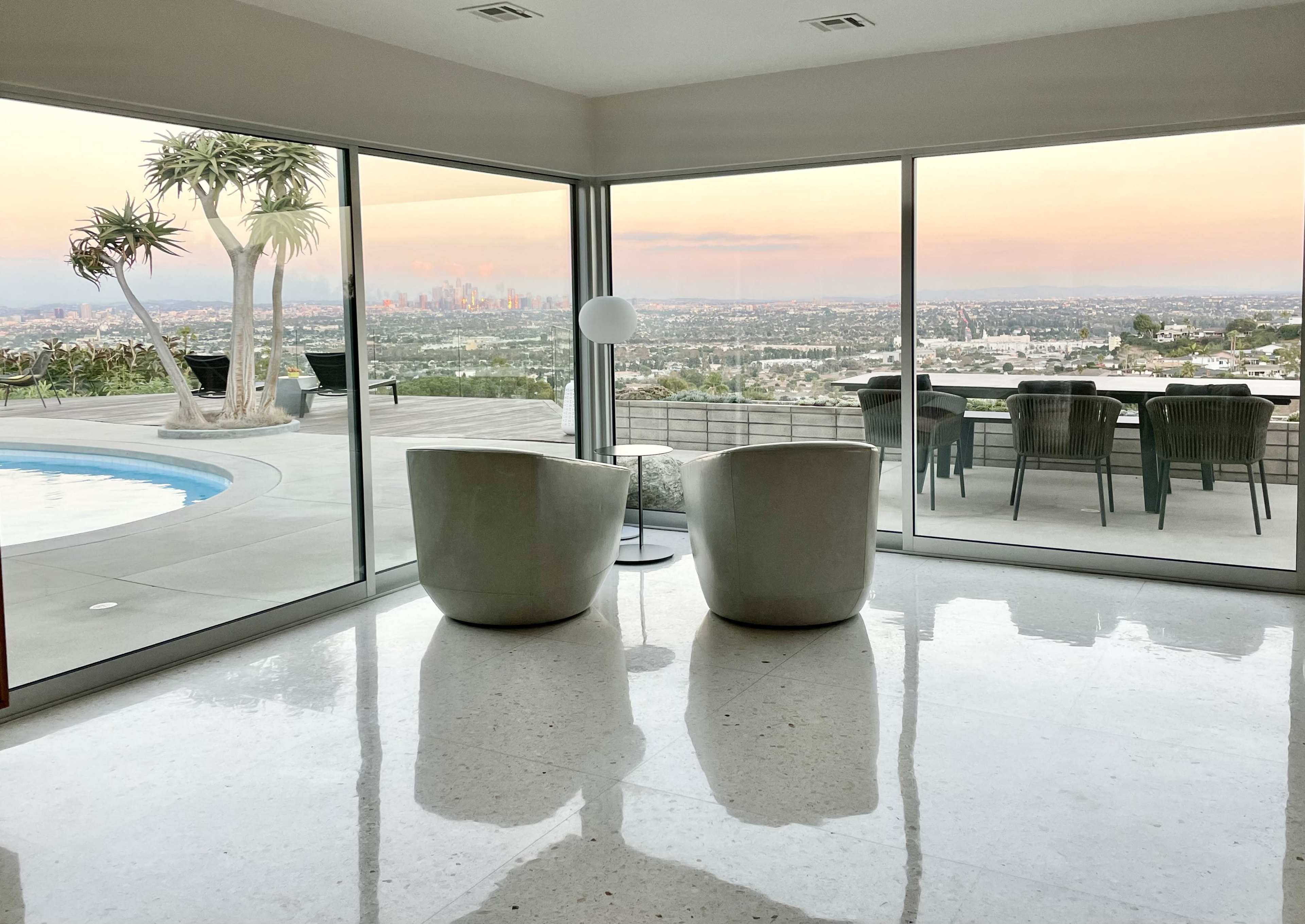 A modern interior with two white chairs facing large glass windows that overlook a city skyline at sunset.