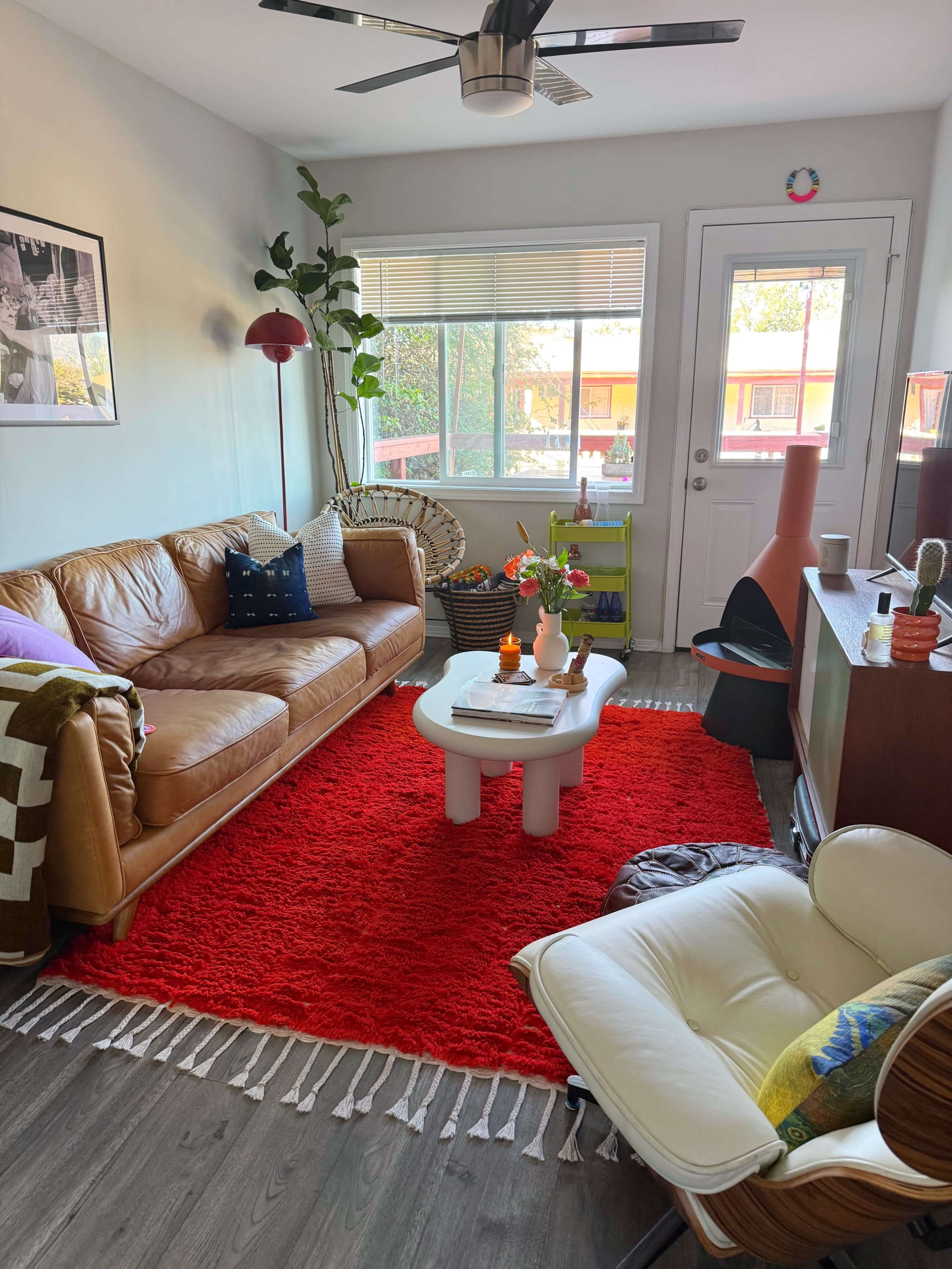The image shows a cozy living room with a brown leather sofa, a red rug, and a white coffee table, complemented by plants and natural light from a nearby window.