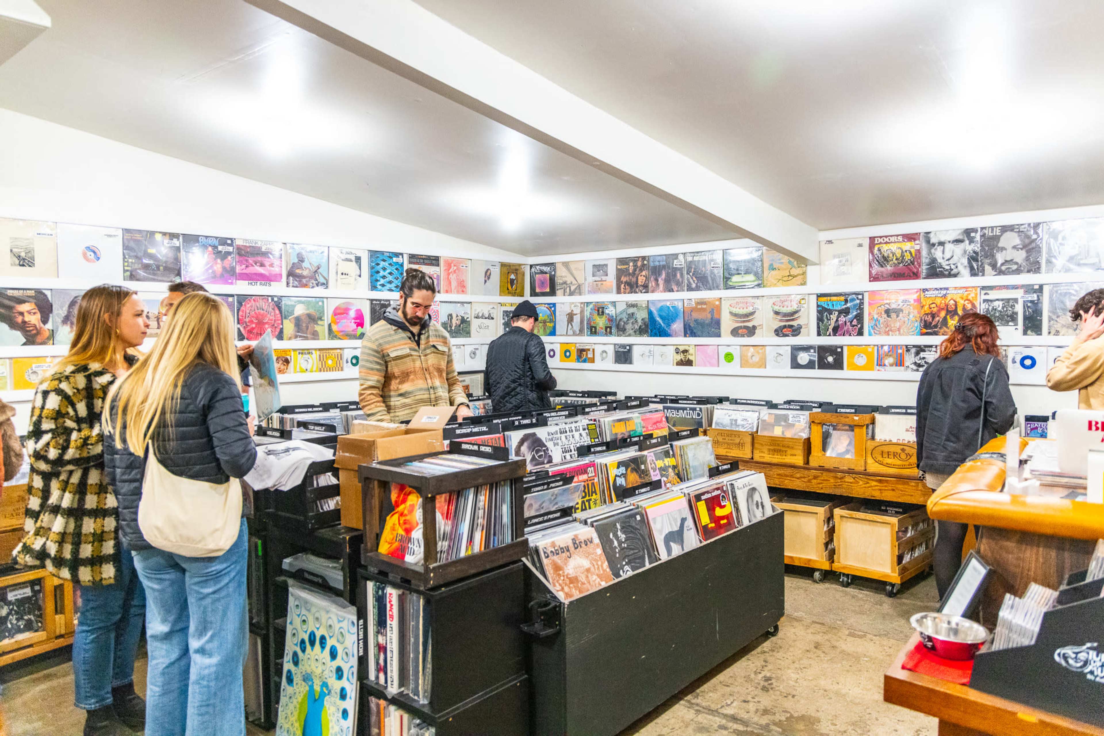 A group of people browse through vinyl records in a music store filled with shelves of albums and displayed art on the walls.