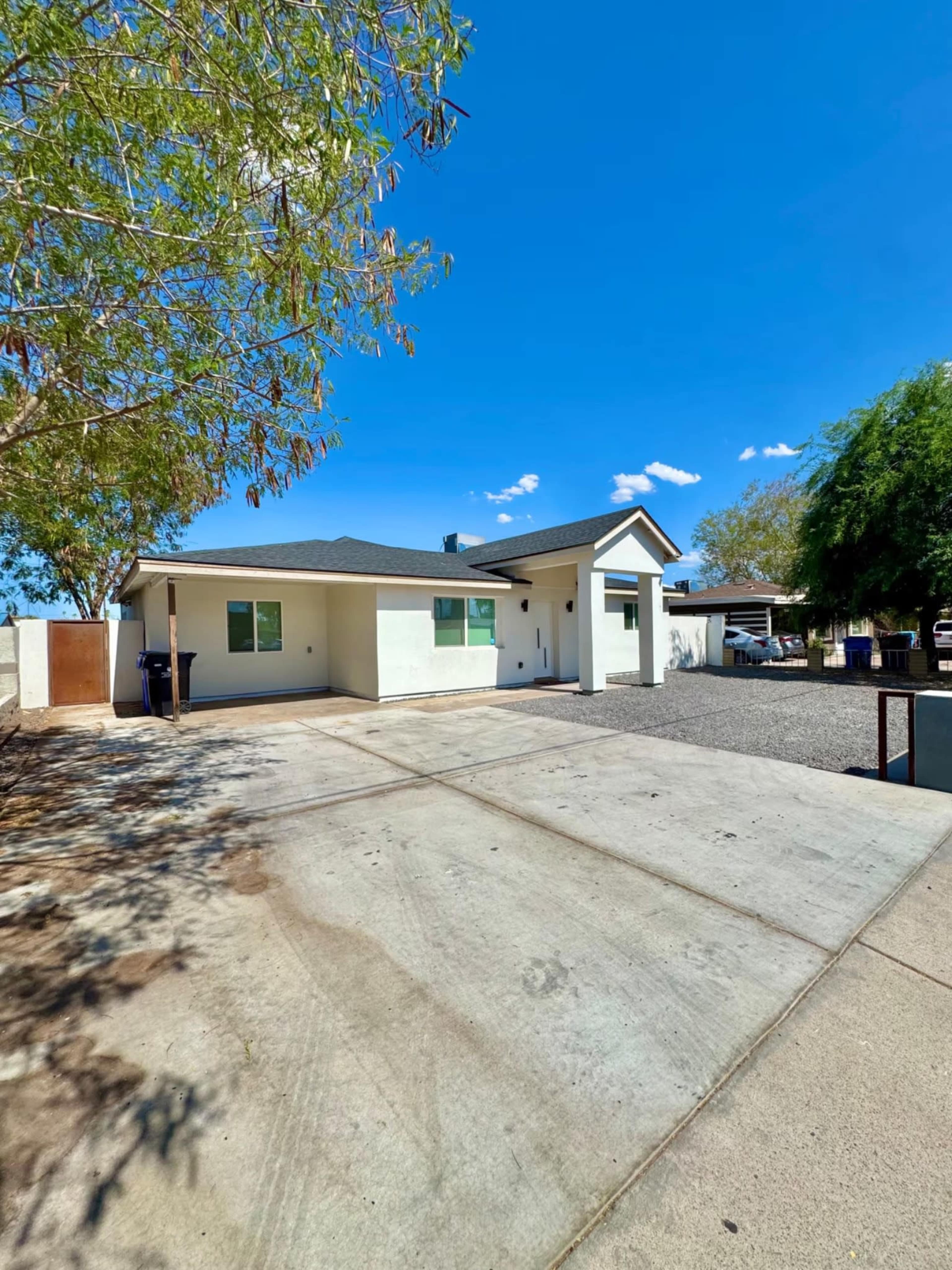 The image shows a single-story house with a light-colored exterior, set on a gravel driveway under a clear blue sky.