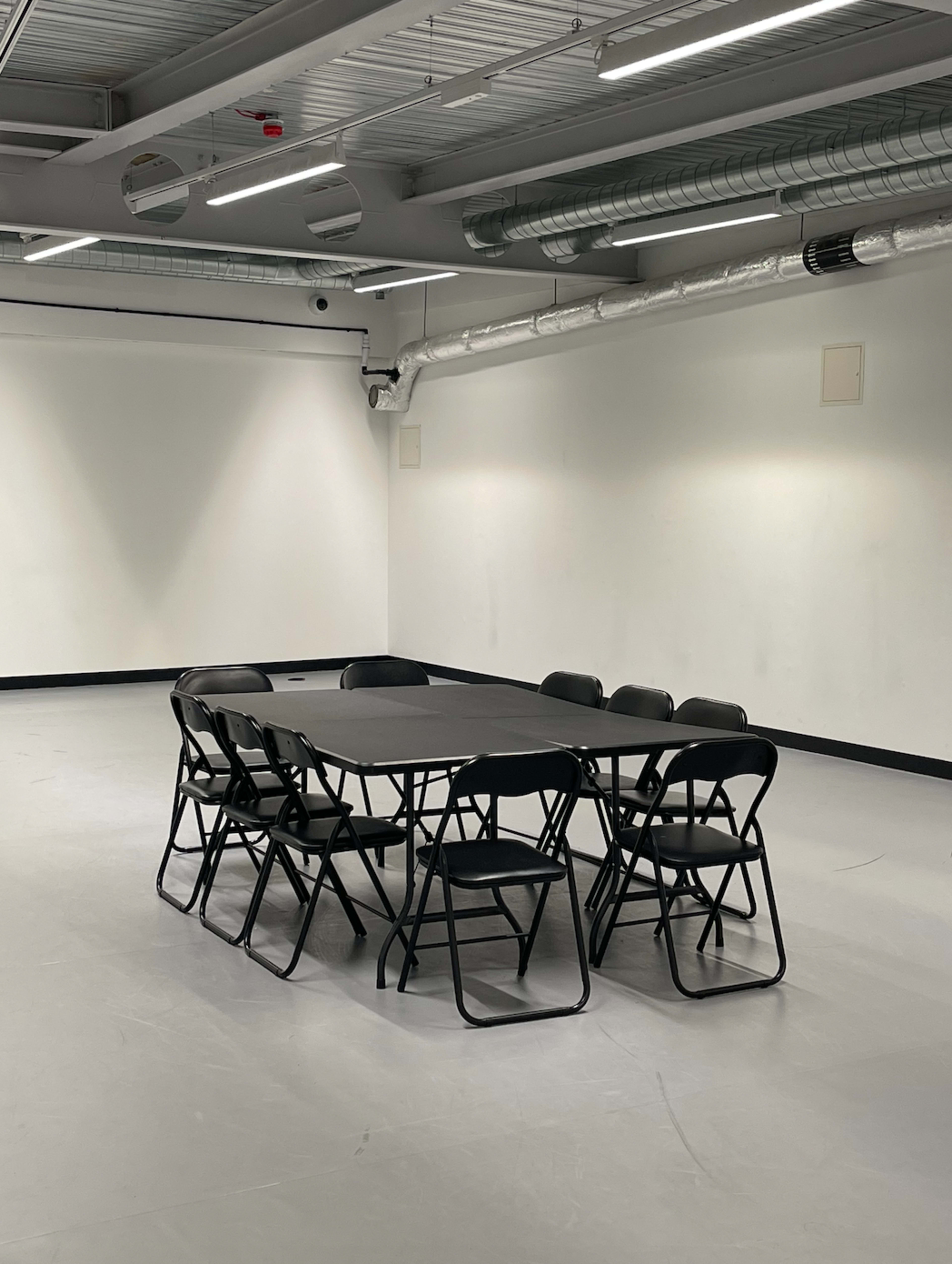 A rectangular table surrounded by eight black folding chairs is positioned in the center of an empty, brightly lit room.