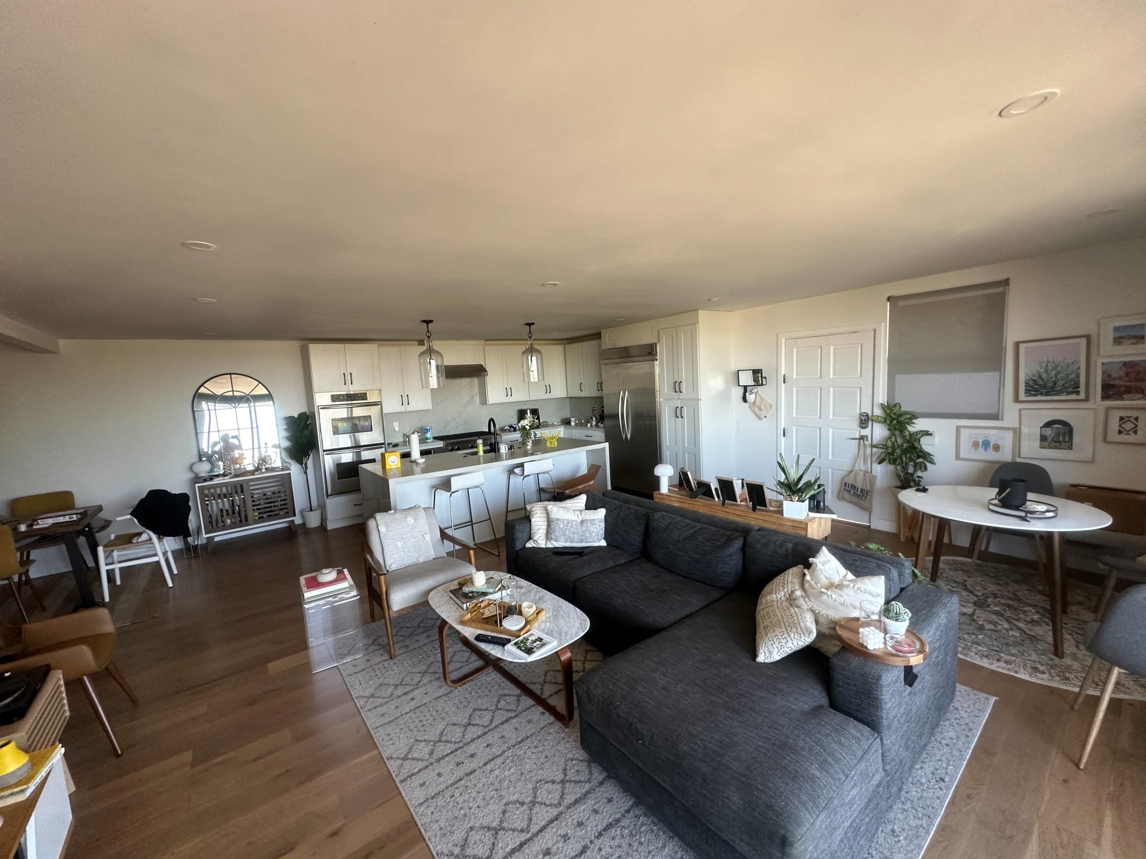 The image shows a modern living space featuring a gray sectional sofa, a dining area with a round table, and a kitchen with stainless steel appliances.