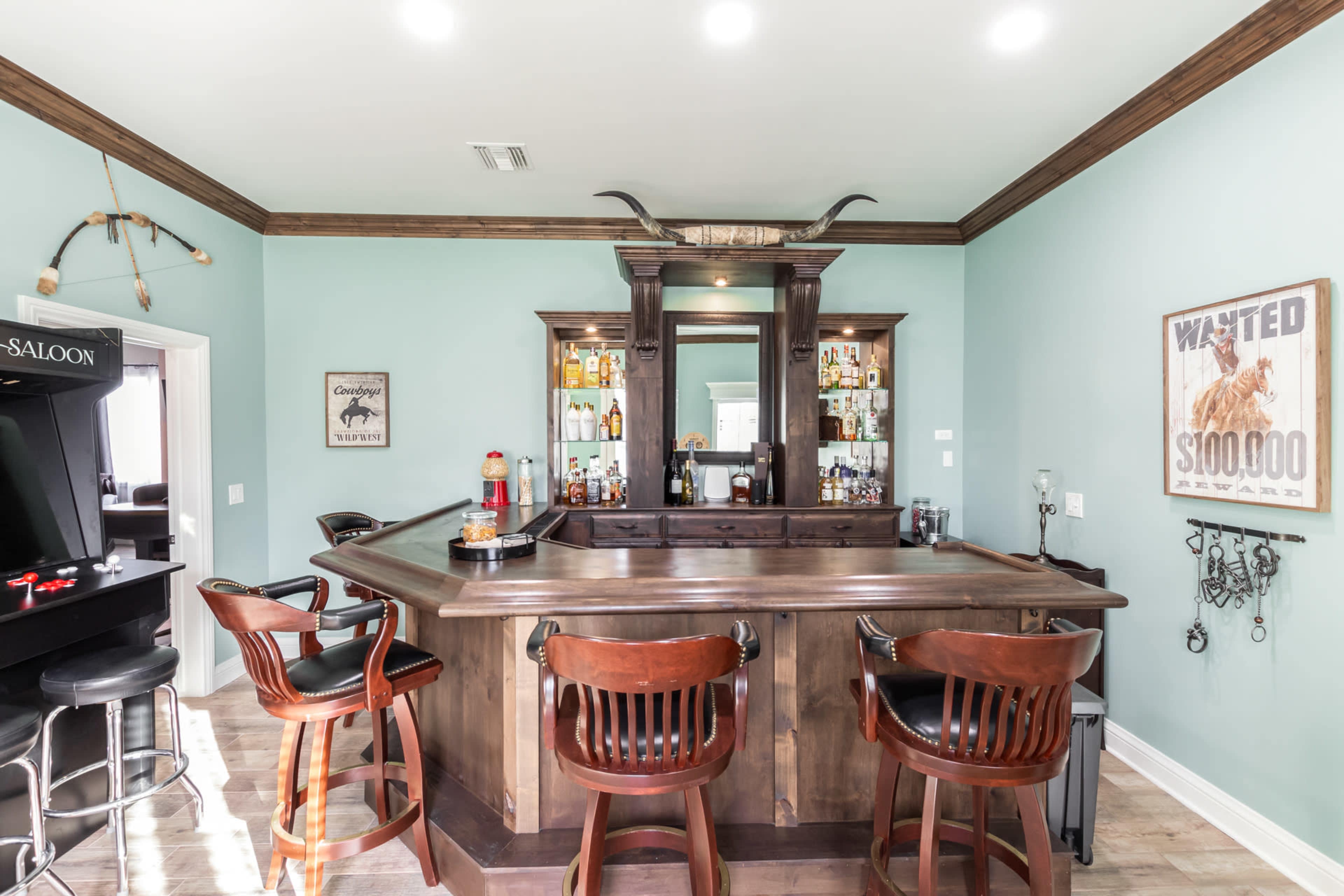 A wooden bar with high stools is surrounded by light blue walls and features shelves of liquor and decor items.