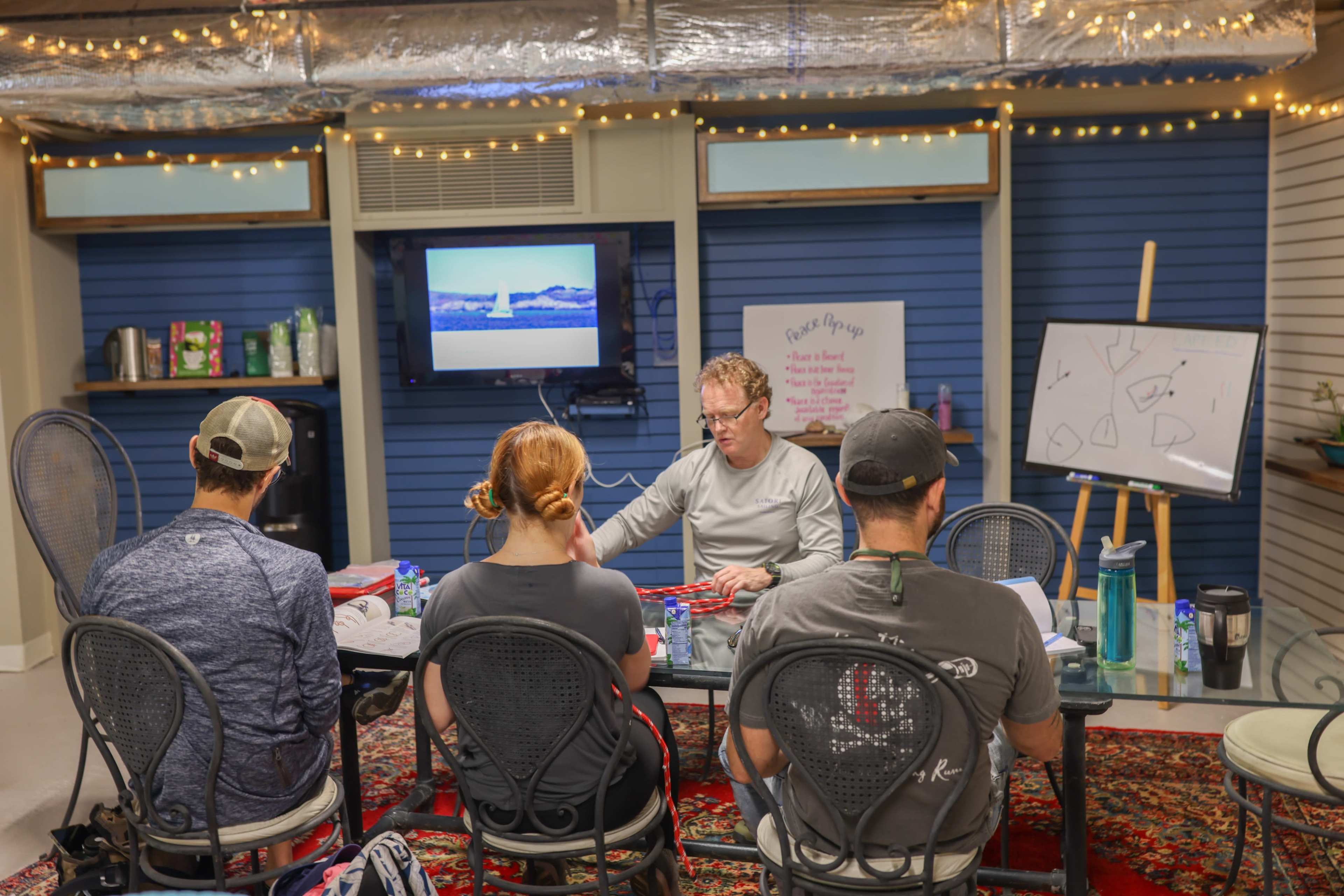A group of four people sits around a table in a room with blue walls and a presentation screen, as one individual leads a discussion or demonstration.