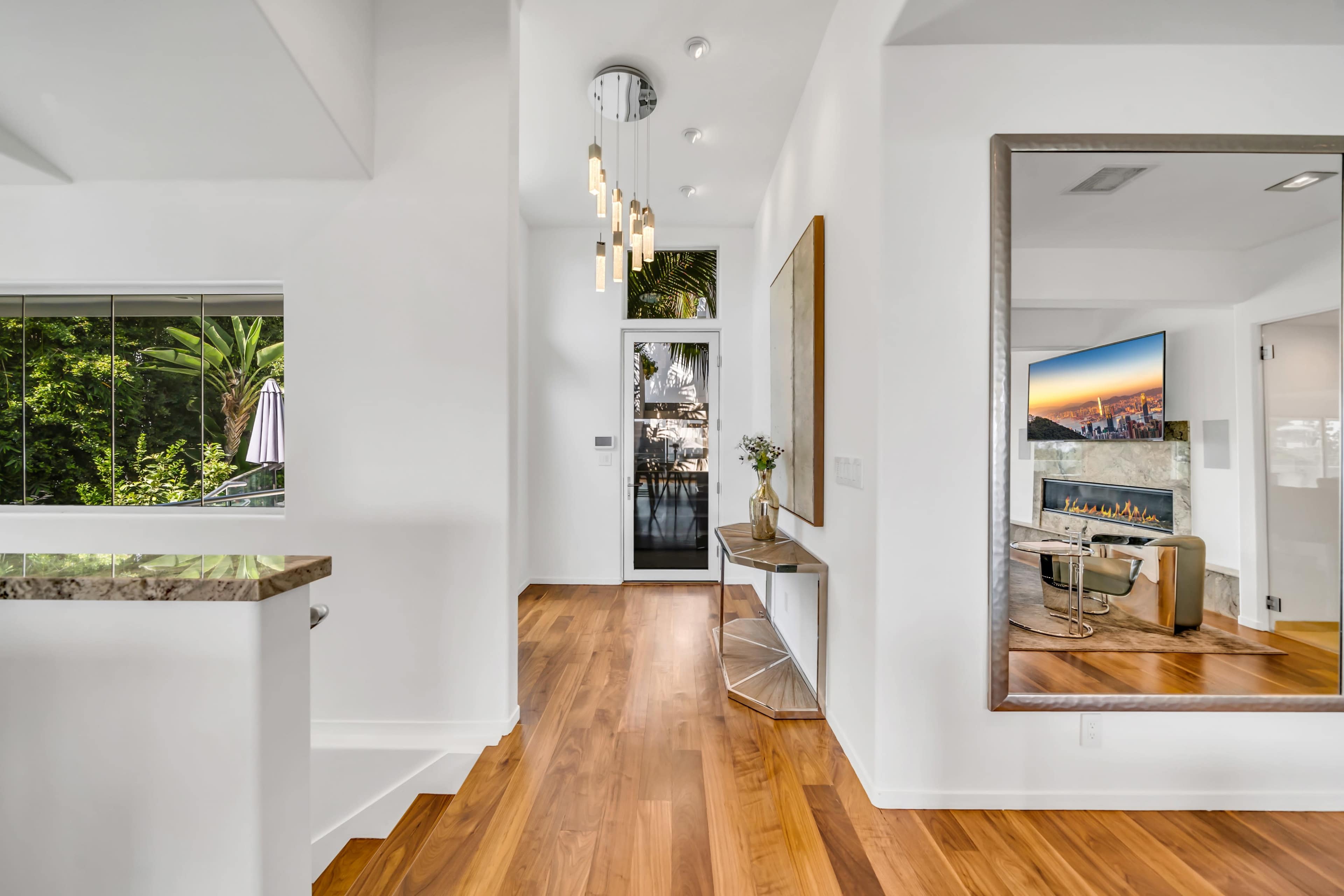The image shows a bright, modern hallway with wooden flooring, a large mirror, and a view of greenery through a window.