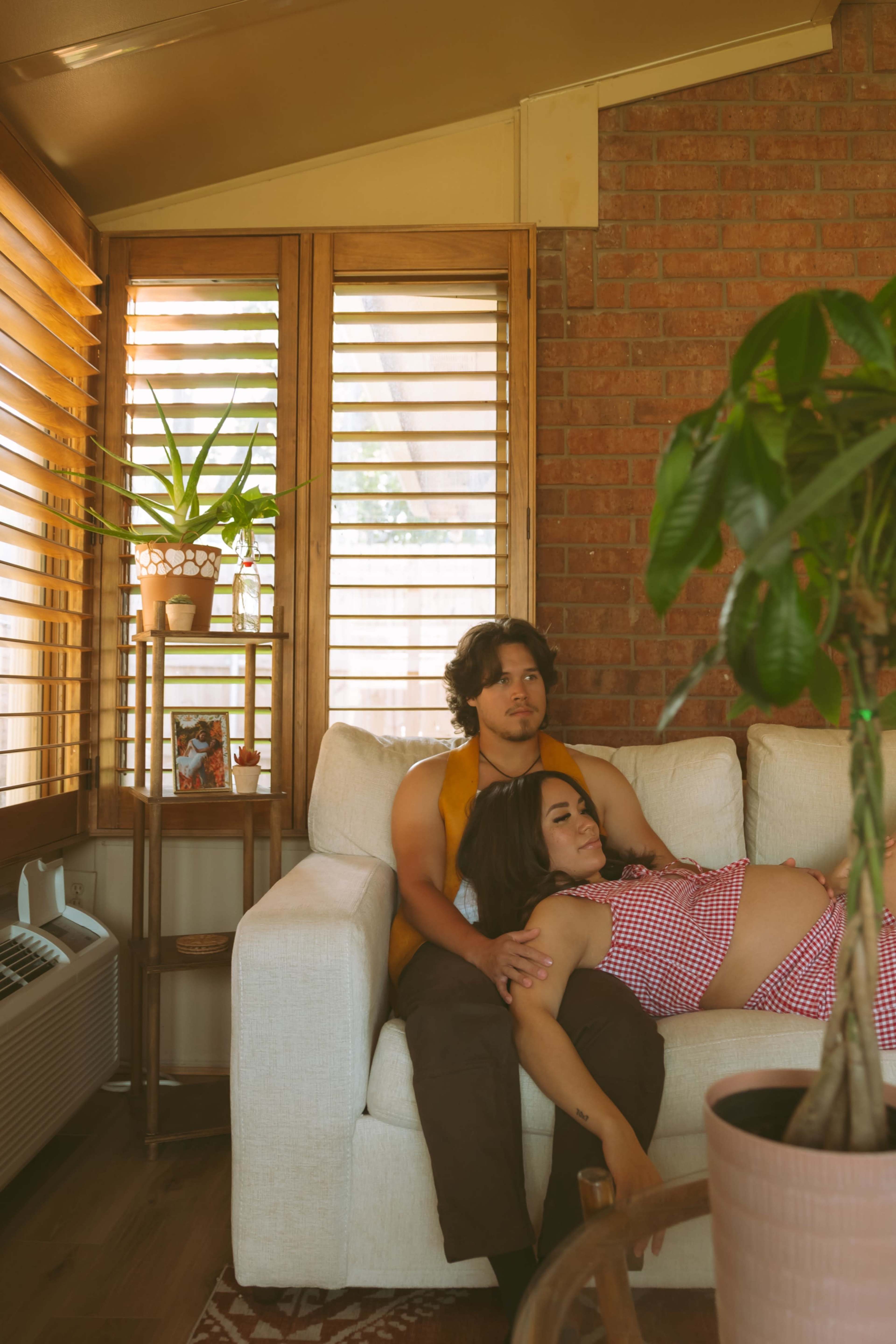 A man sits on a couch while a woman reclines against him in a room with wooden blinds and plants.