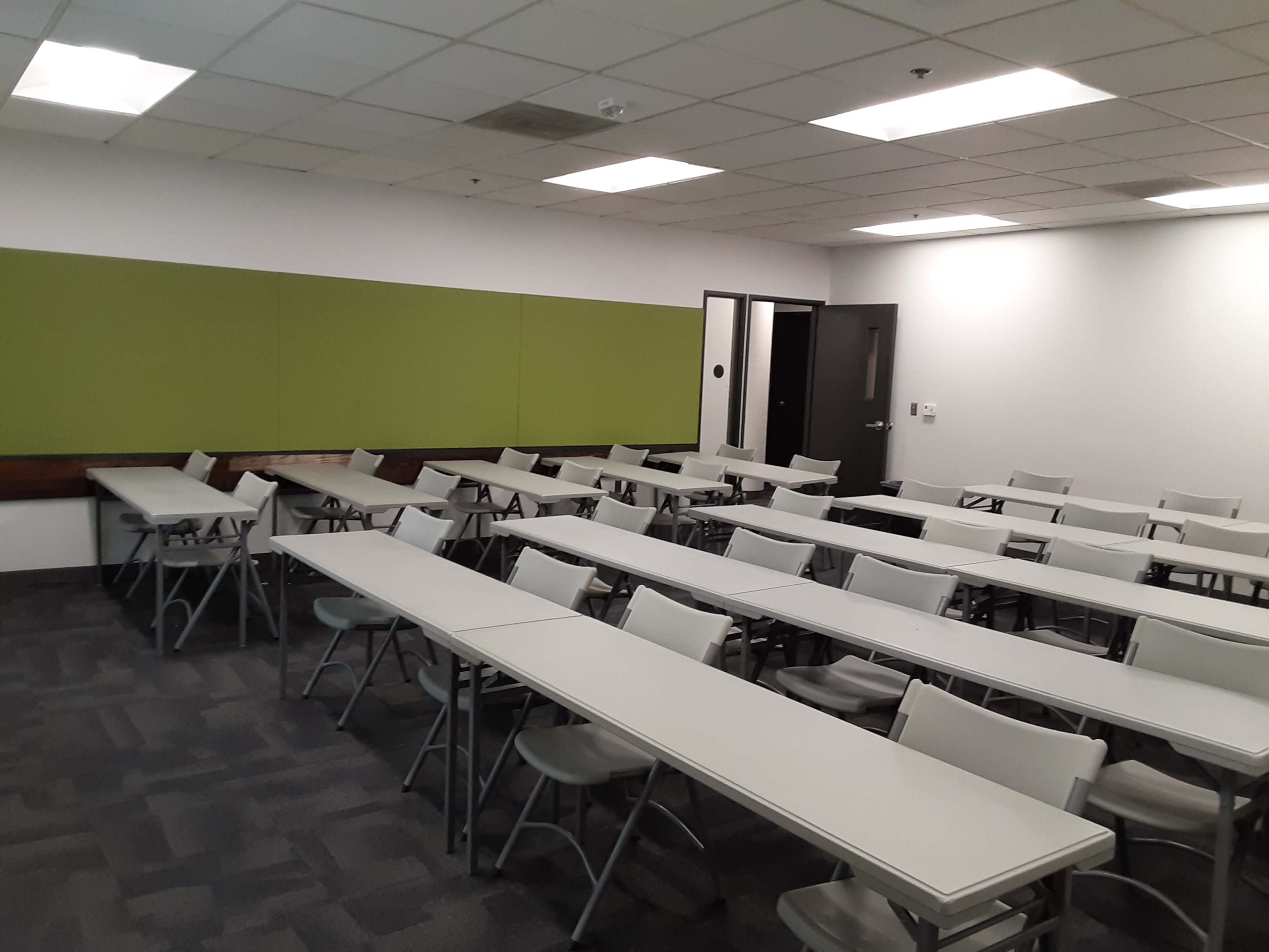 A classroom featuring multiple rows of tables and chairs arranged in a rectangular layout, with a green wall section in the background.
