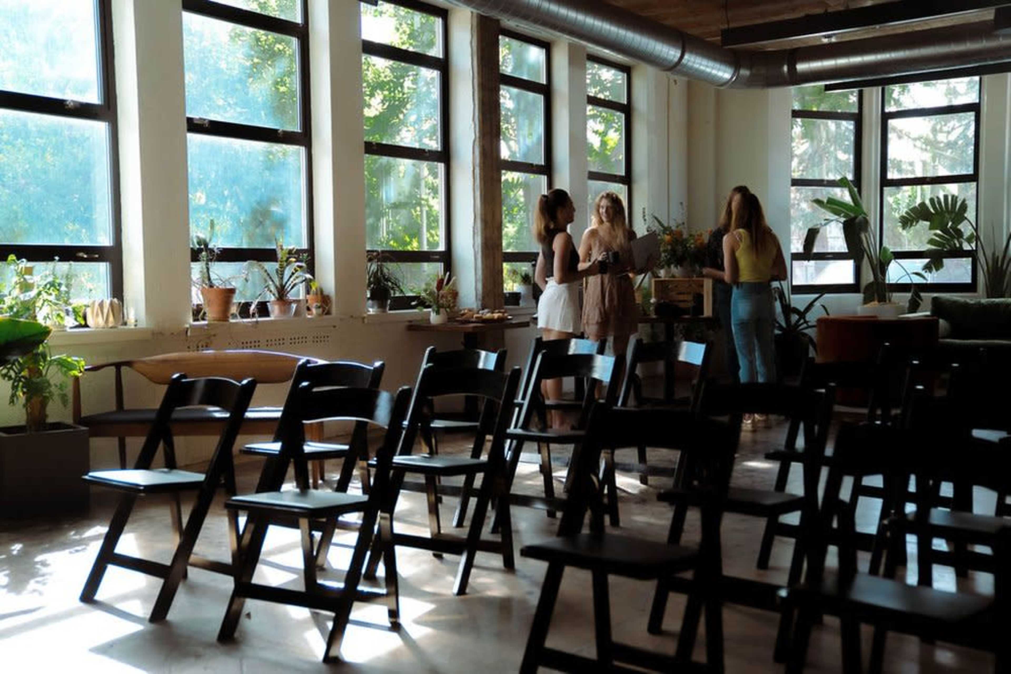 Three women are standing and conversing in a brightly lit room with large windows and empty folding chairs arranged in rows.