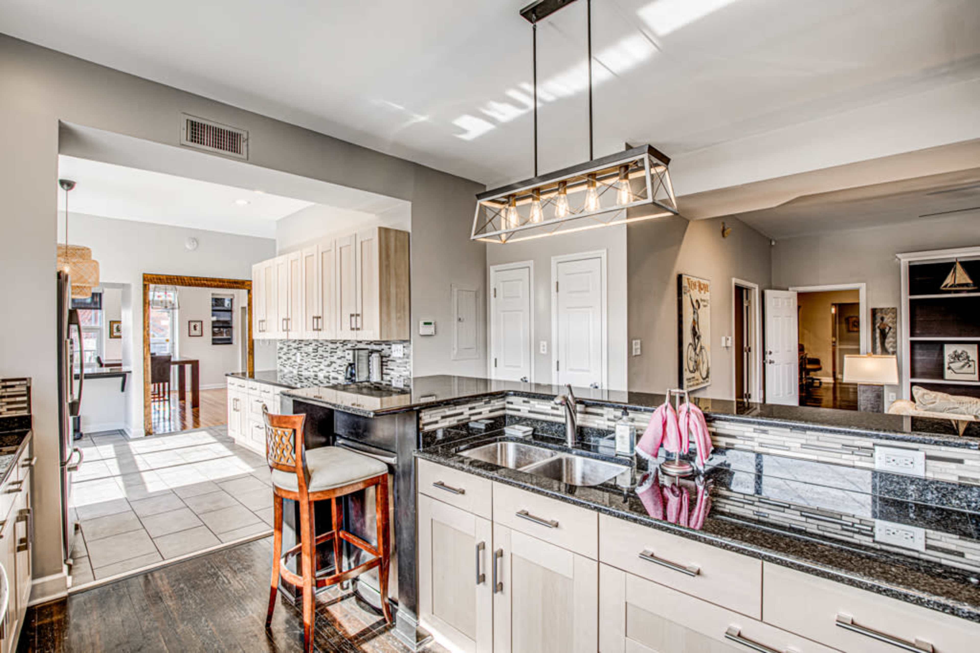 A modern kitchen features a central island with a granite countertop, light fixtures, and a bar stool, with cabinets and a tiled floor.