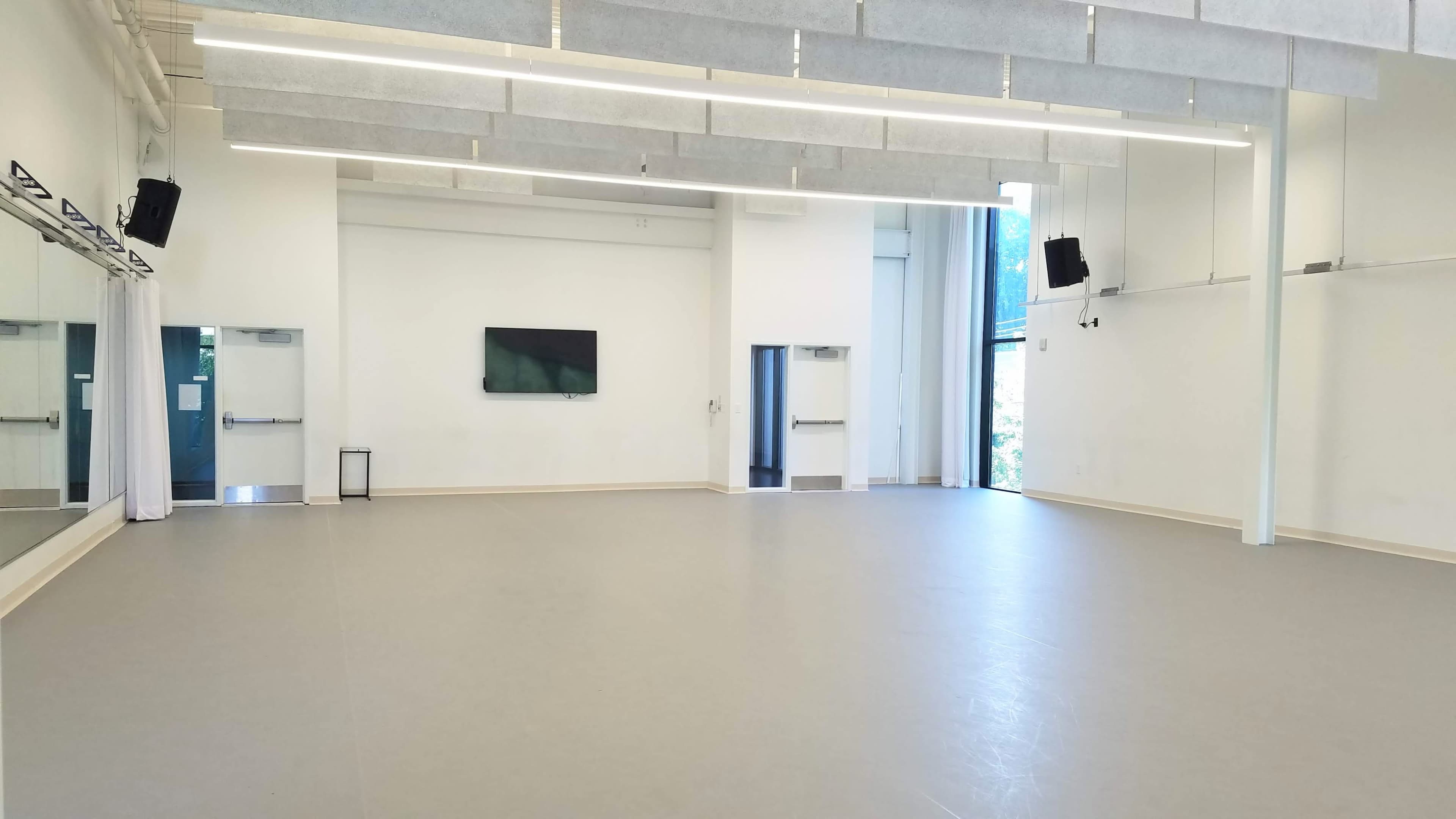 The image shows a spacious, well-lit dance studio with large windows and a wooden floor, featuring mirrors on one wall and minimal furnishings.