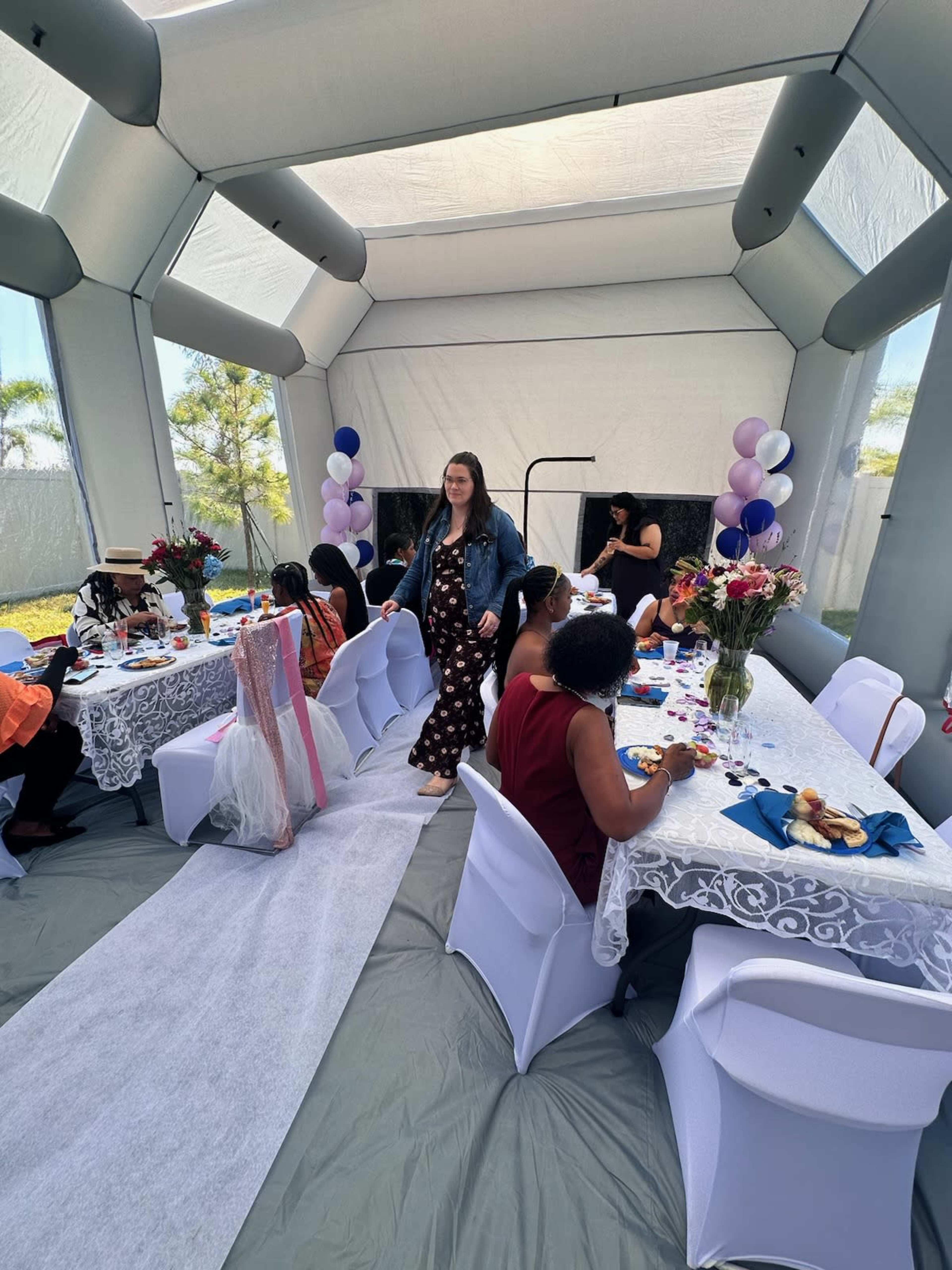 A woman in a denim jacket walks through a decorated tent where a small group of people are seated at tables, enjoying food and drinks.