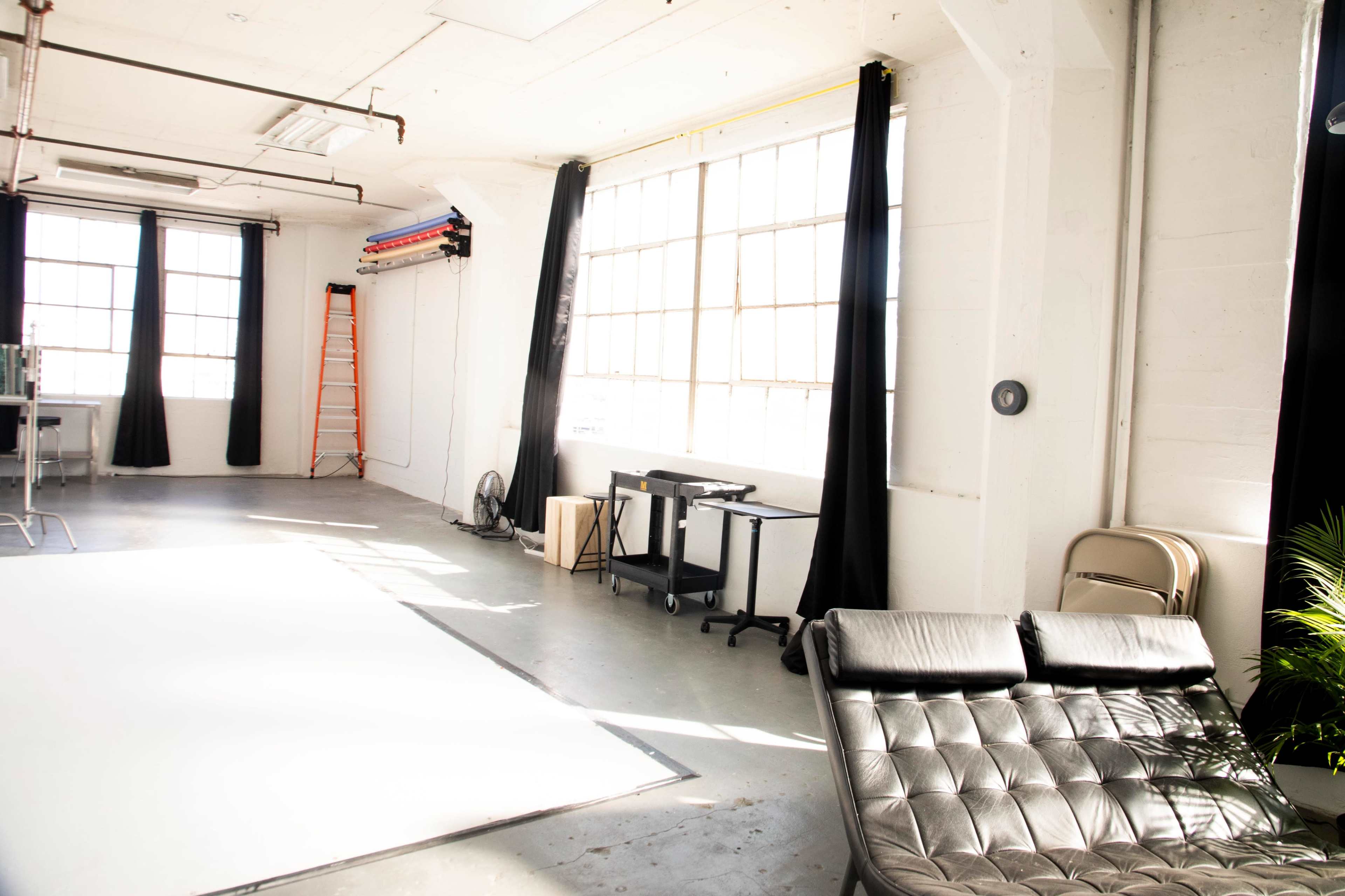 A bright, spacious studio with large windows, a black chaise lounge, a desk, and a ladder against the wall.