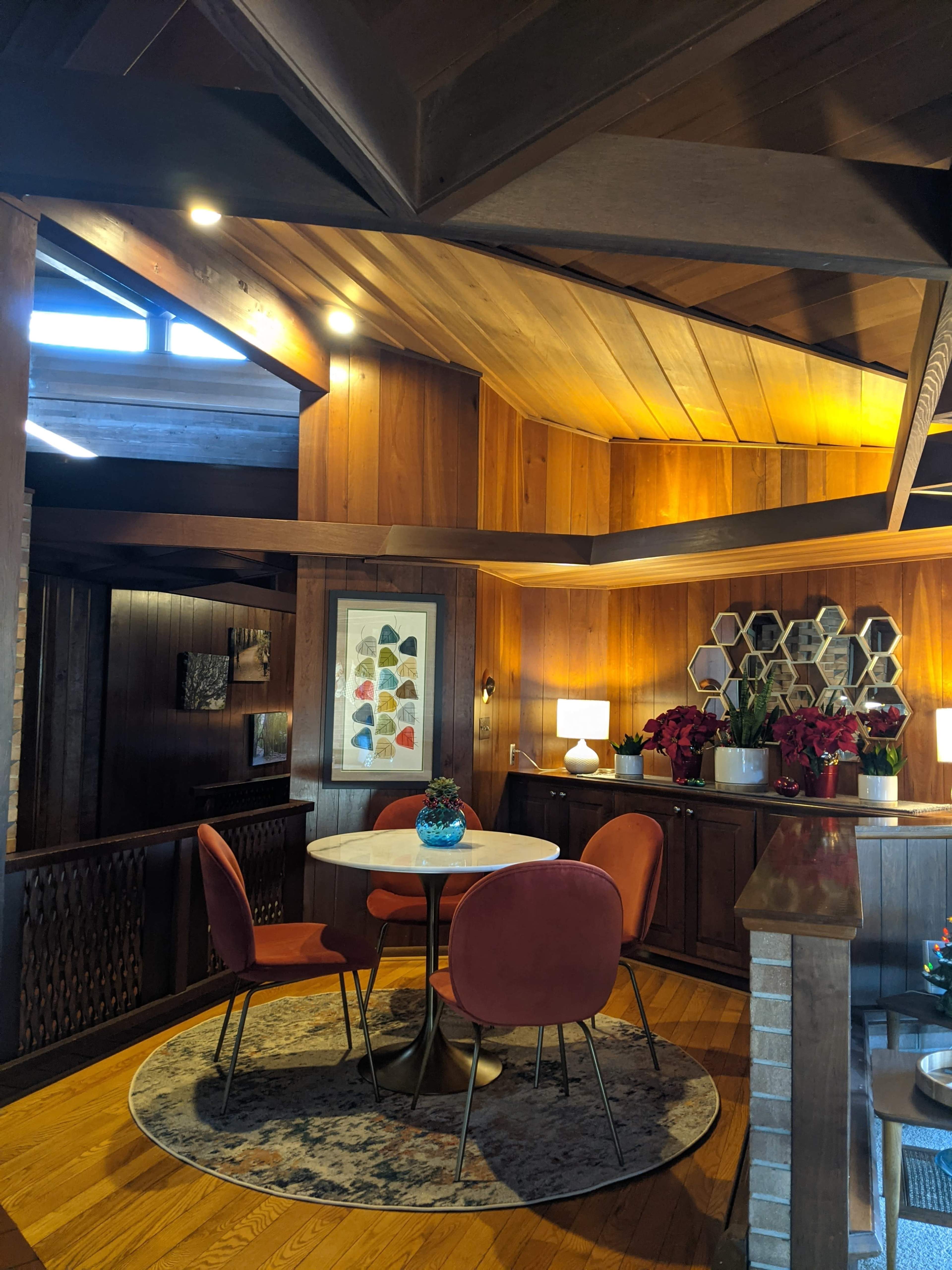 A well-lit dining area with a round table surrounded by four orange chairs, set against wooden walls and a decorative rug.
