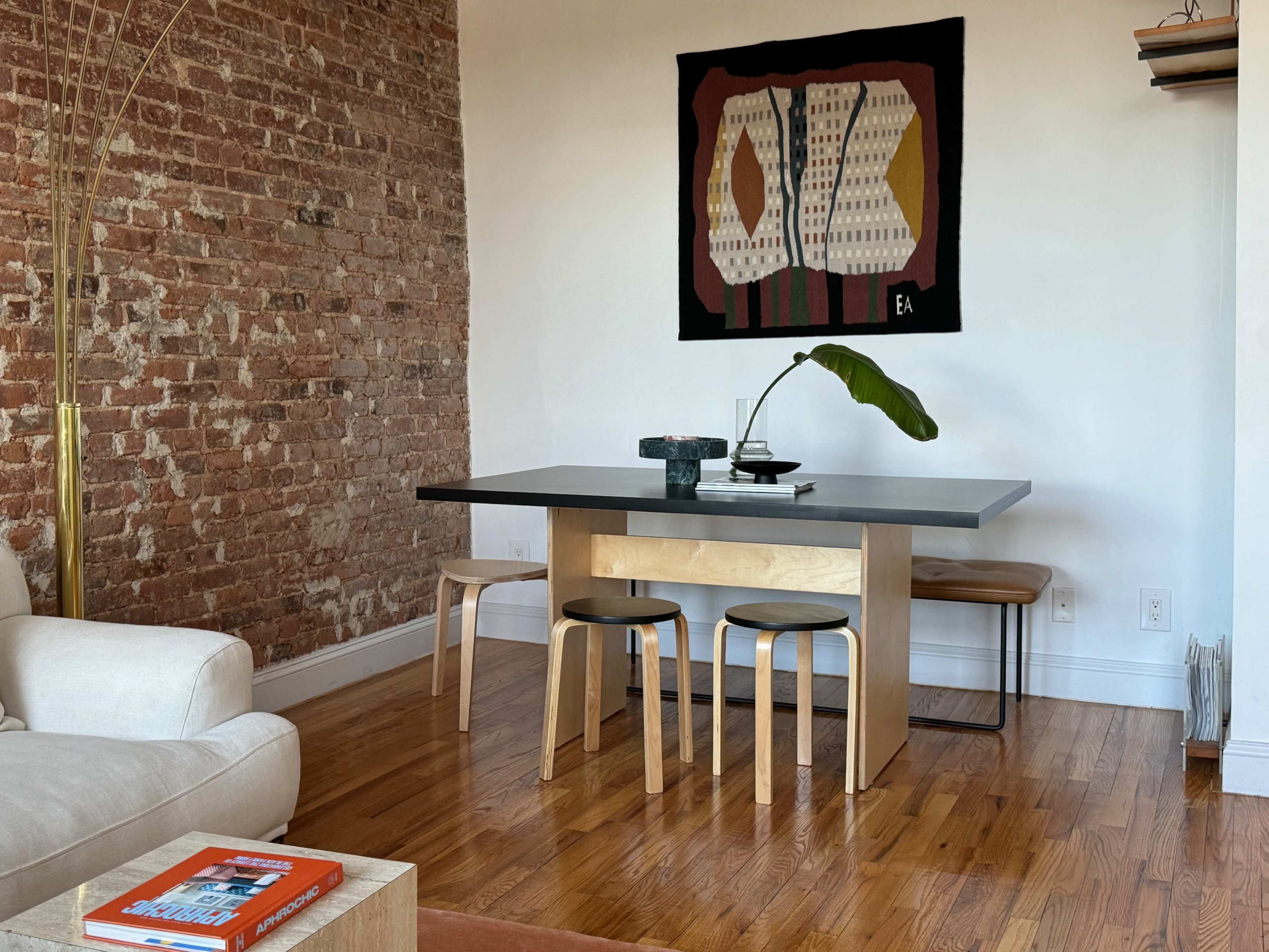 A modern dining area features a wooden table with two stools, a vase with a plant, and a textured wall hanging above the table, all set against a backdrop of exposed brick.