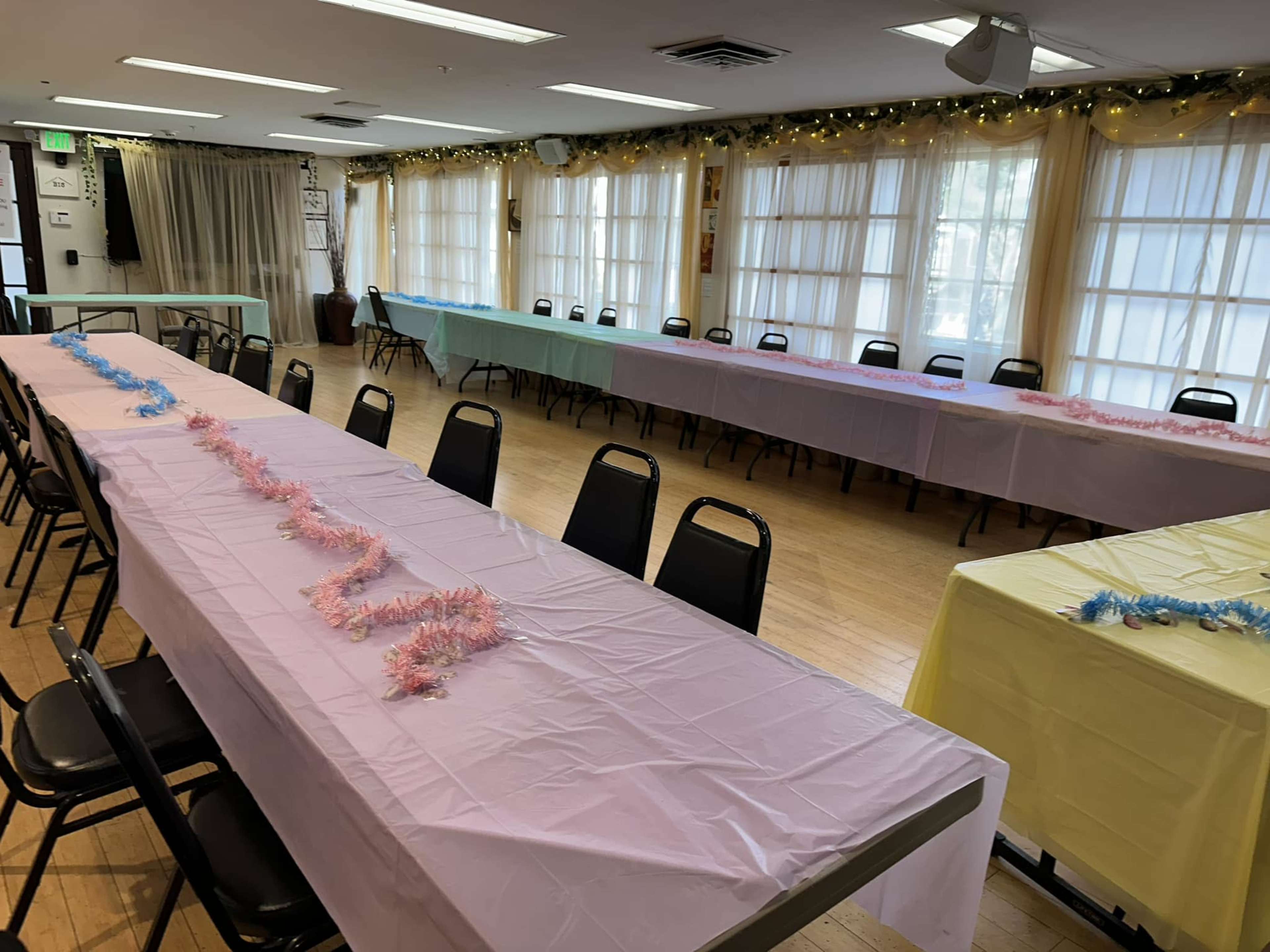 The image shows a large room with several long tables covered in pastel-colored tablecloths, arranged with black chairs, and decorated with tinsel.
