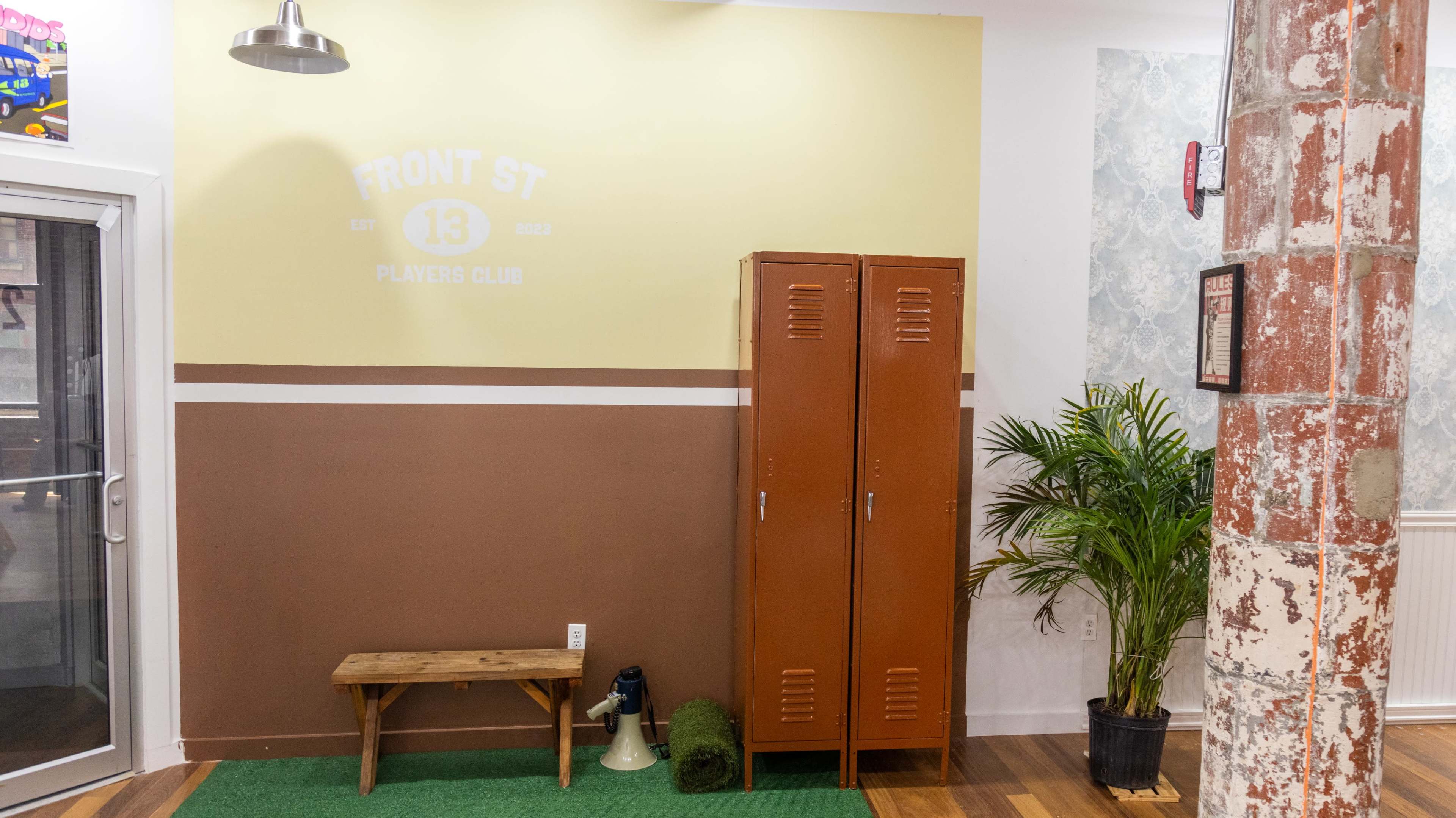A small interior corner featuring a wall with the text "FRONT ST 13 PLAYERS CLUB," a brown bench, a set of orange lockers, and a potted plant.