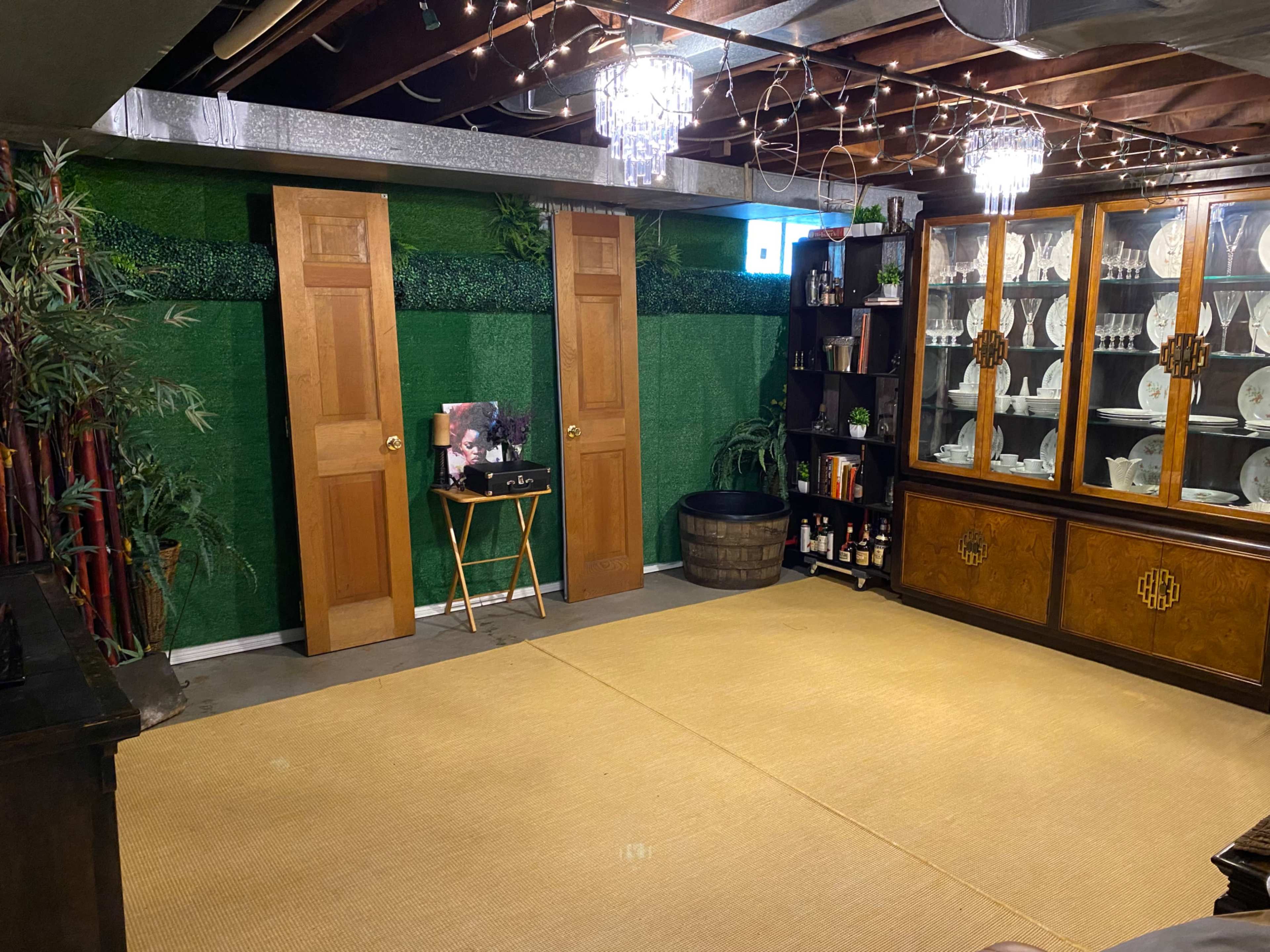 The image shows a spacious basement room with a bamboo-themed decor, two wooden doors, a glass-front cabinet displaying dinnerware, and a clear floor mat.