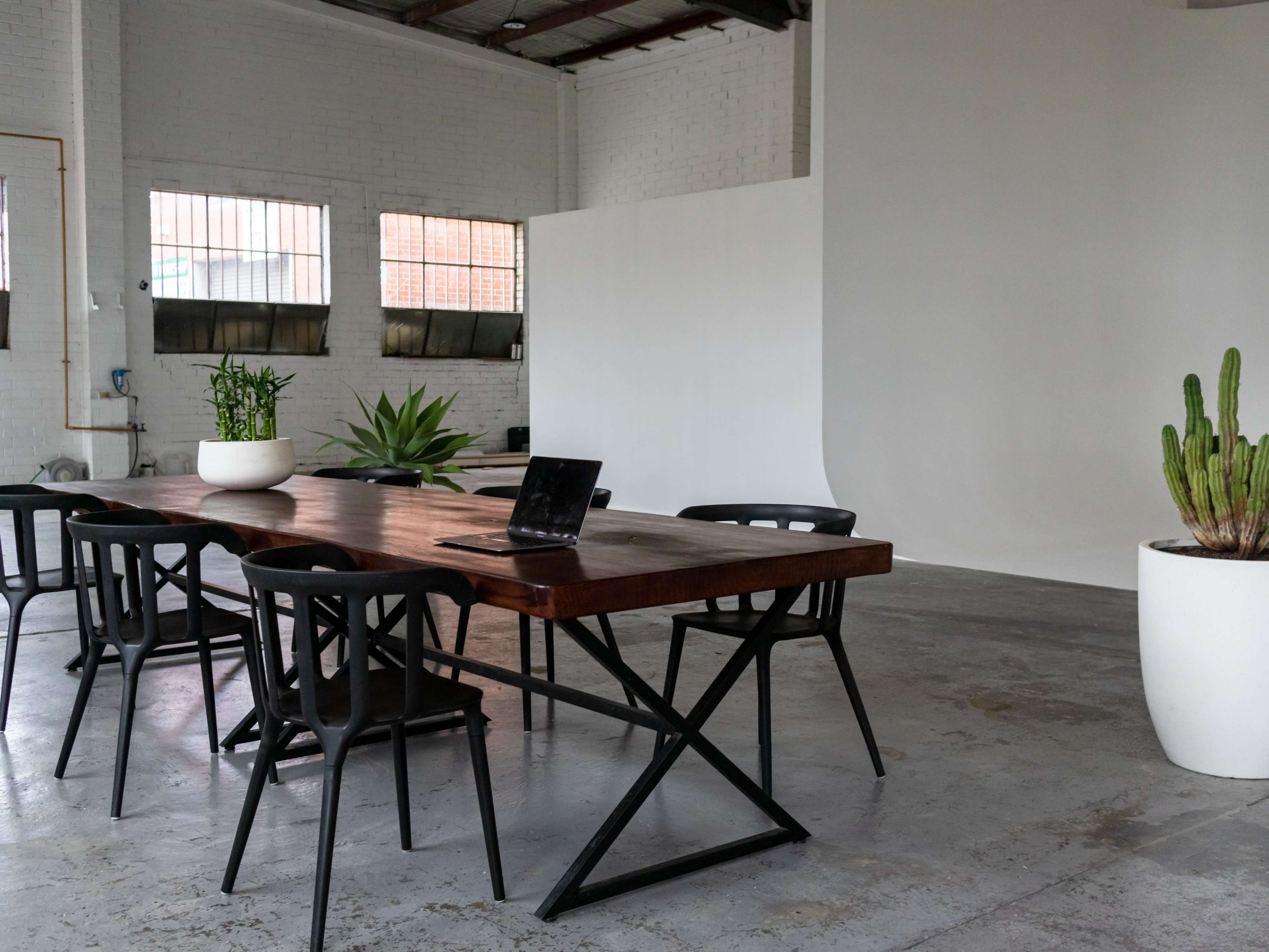 A modern conference room features a wooden table surrounded by black chairs, with a laptop positioned on the table and potted plants in the background.