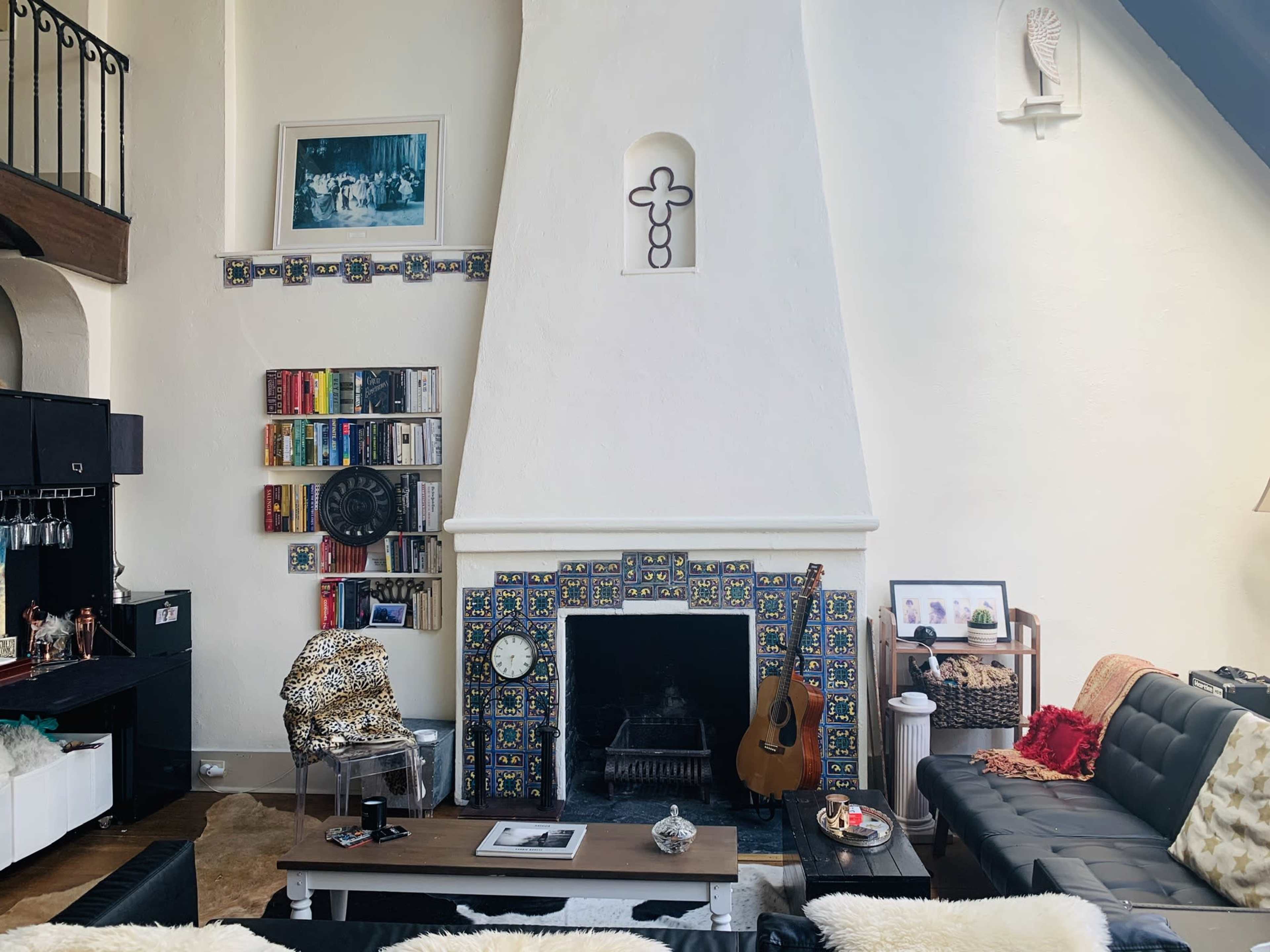 A living room with a large white fireplace adorned with colorful tiles, surrounded by bookshelves and a guitar leaning against the wall.