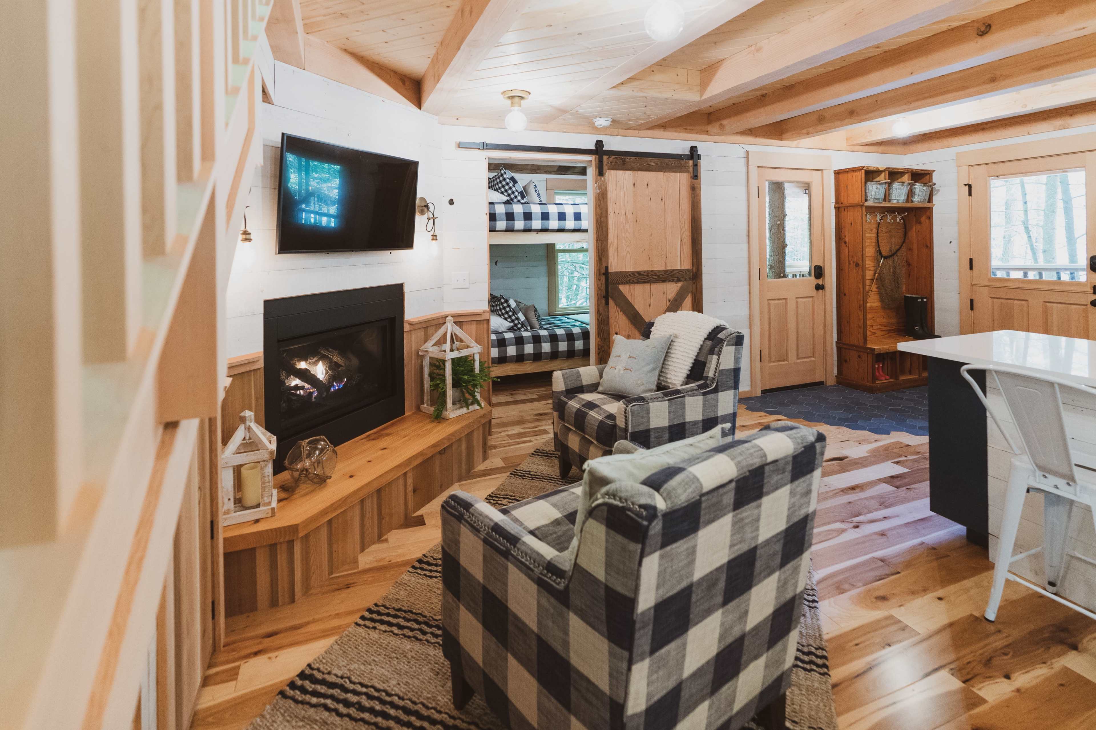 The interior of a cozy cabin features a fireplace, two patterned armchairs, a bunk bed, and a dining area with a modern table.