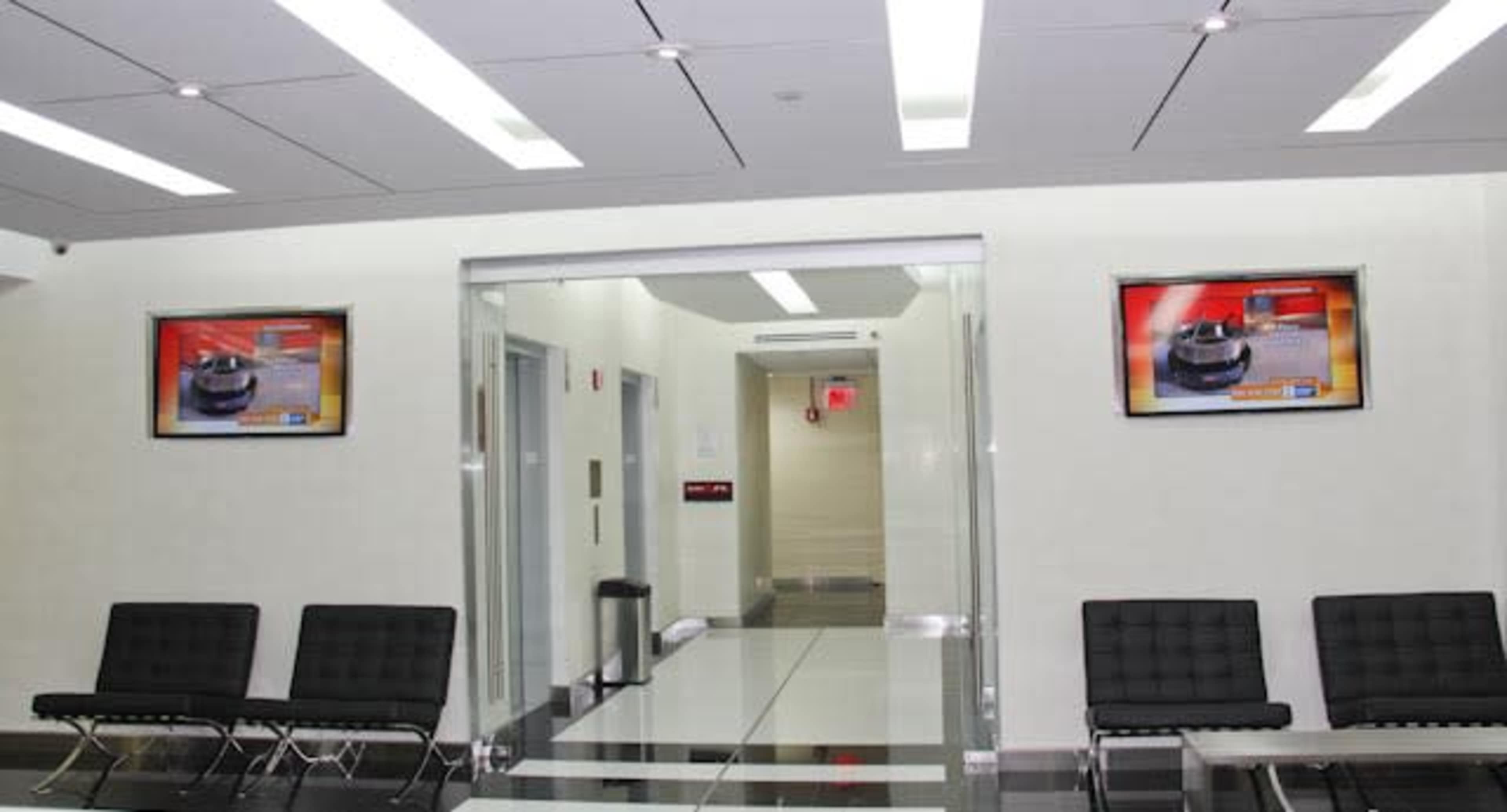The image shows a modern waiting area with black benches, two televisions mounted on the wall, and a glass door leading to another hallway.