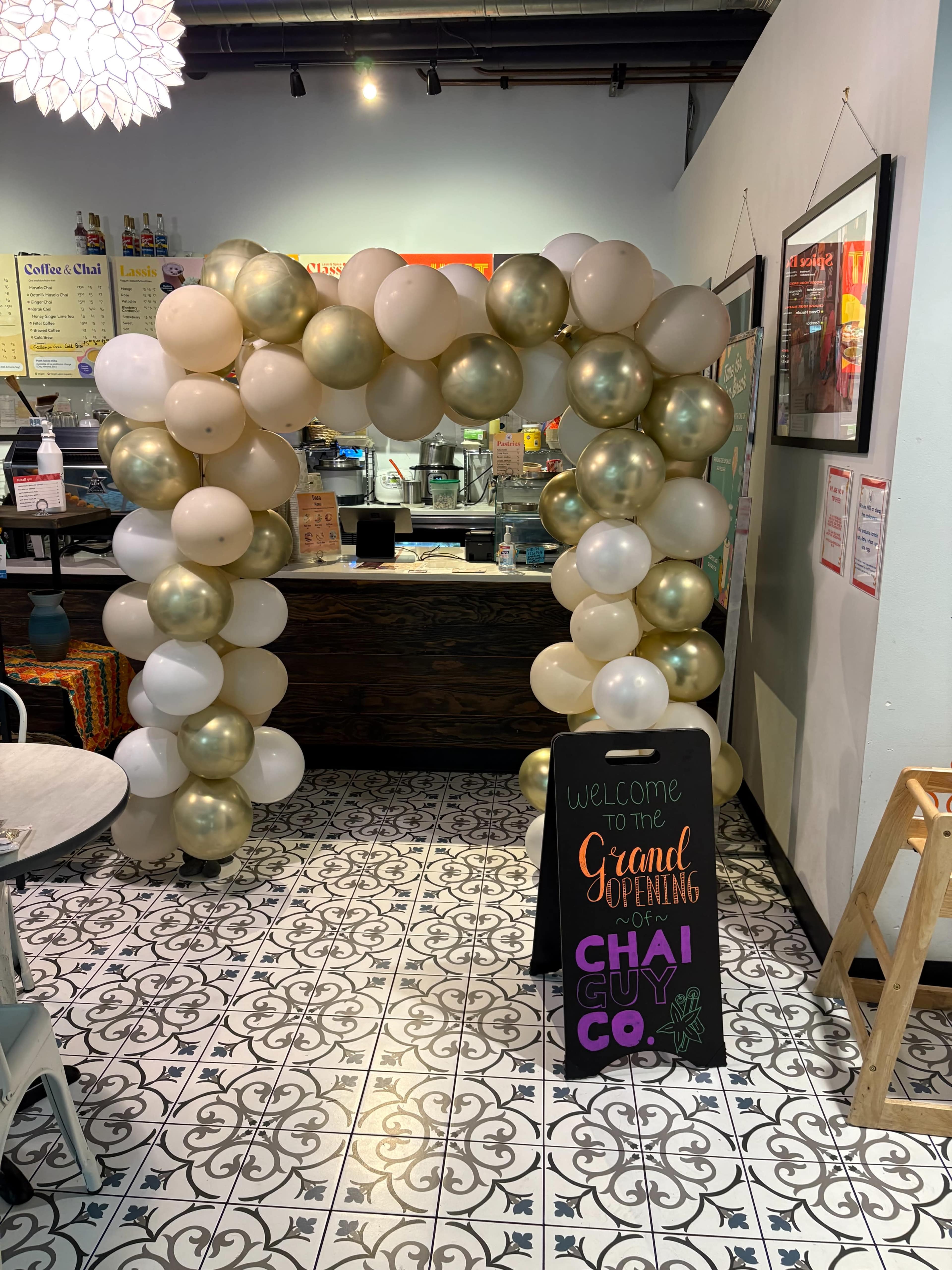 A decorative arch made of white and gold balloons stands at the entrance of a cafe, where a sign announces the grand opening of "Chai Guy Co."