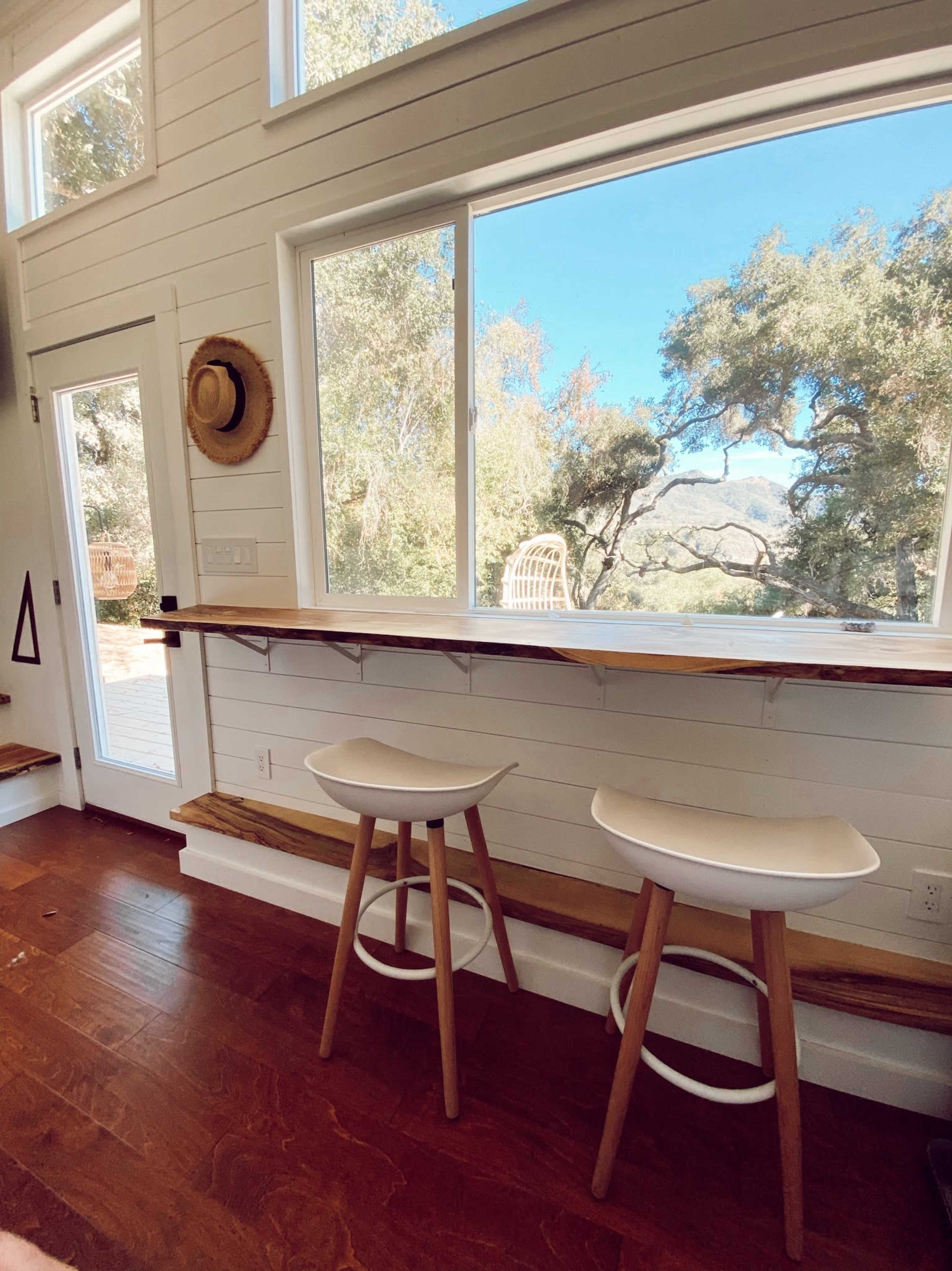 A bright, modern interior with a wooden counter and two white bar stools facing a large window that offers a view of trees and mountains.