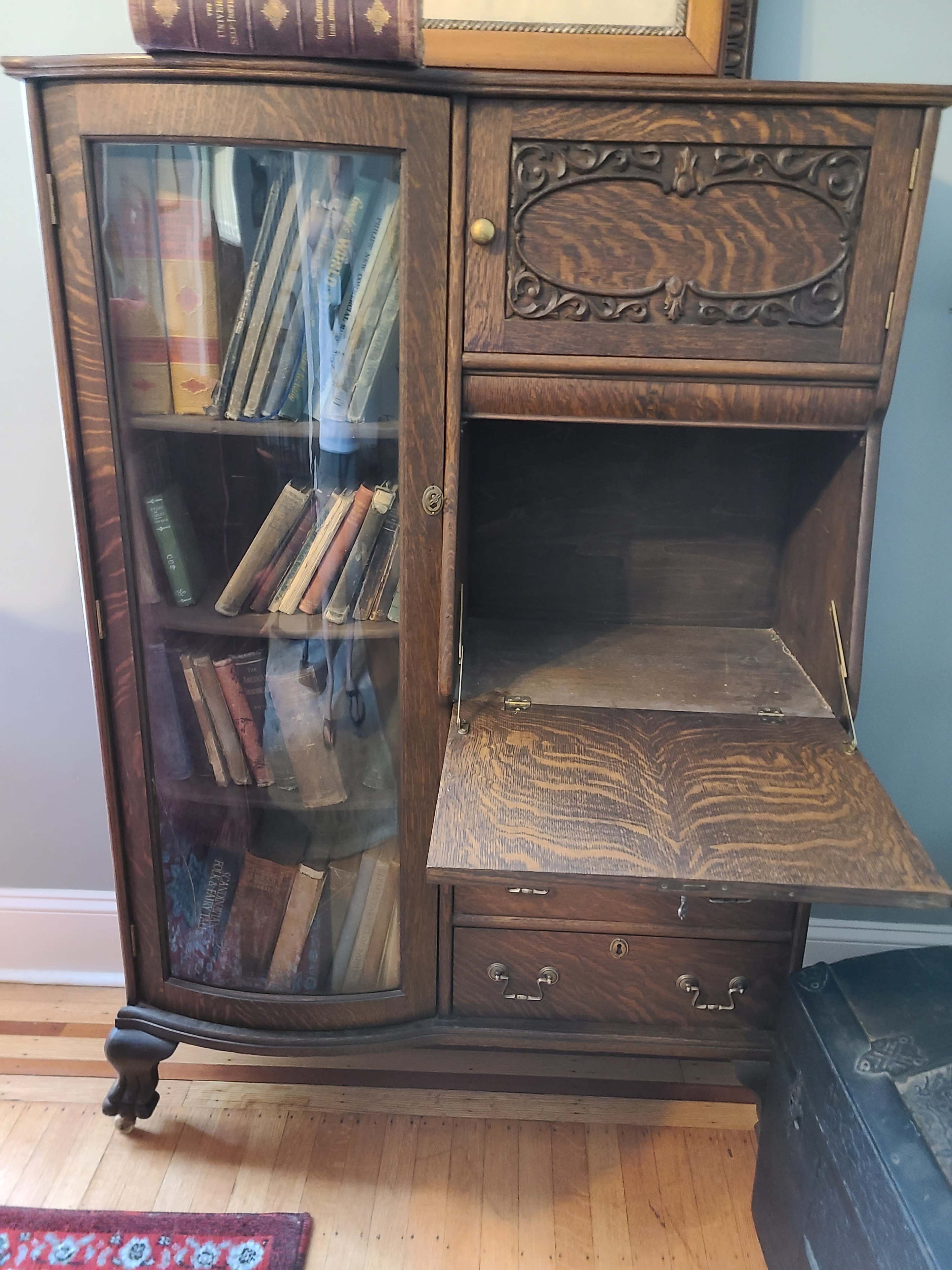 An antique wooden cabinet with a glass door, revealing several old books on its shelves and a lower compartment that is open.