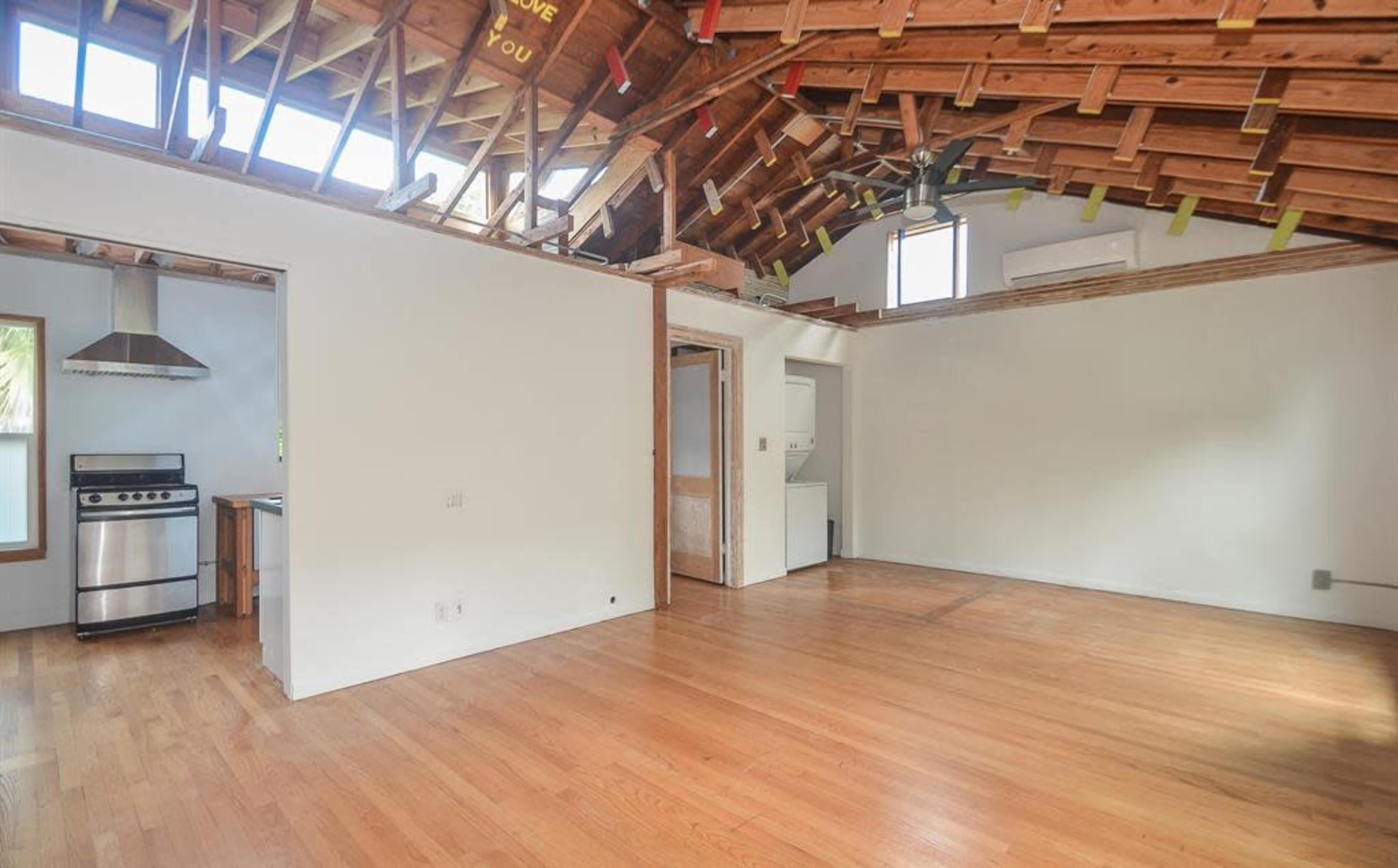 The image shows an interior space with exposed wooden beams, hardwood flooring, and a kitchen area featuring a stainless steel stove and a laundry facility in the corner.