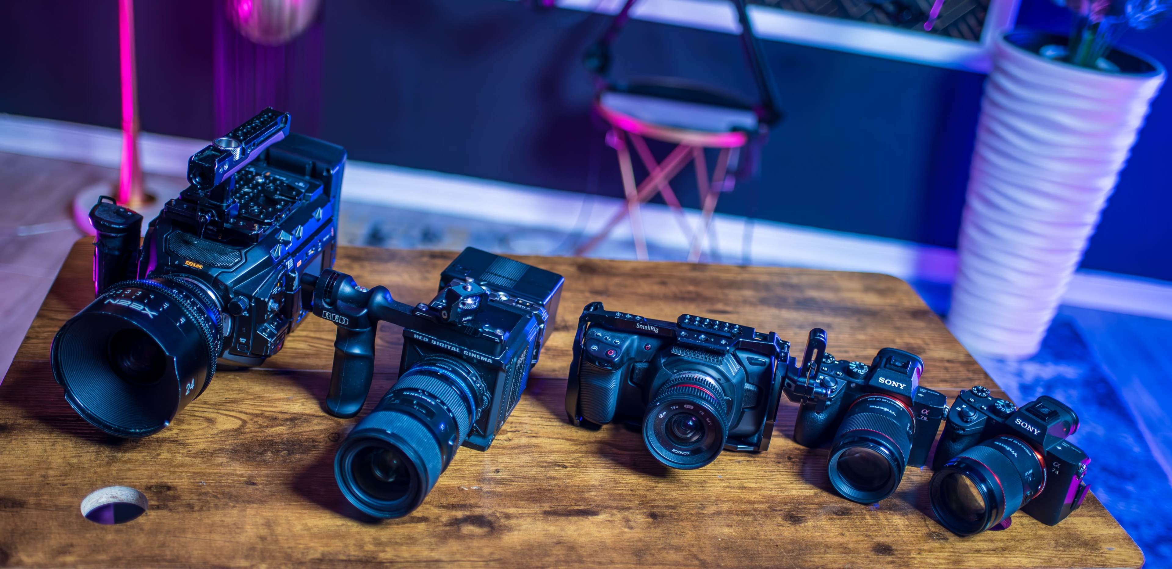A wooden table displays a variety of cameras, including DSLRs and mirrorless models, arranged from left to right in a well-lit room with colorful lighting.