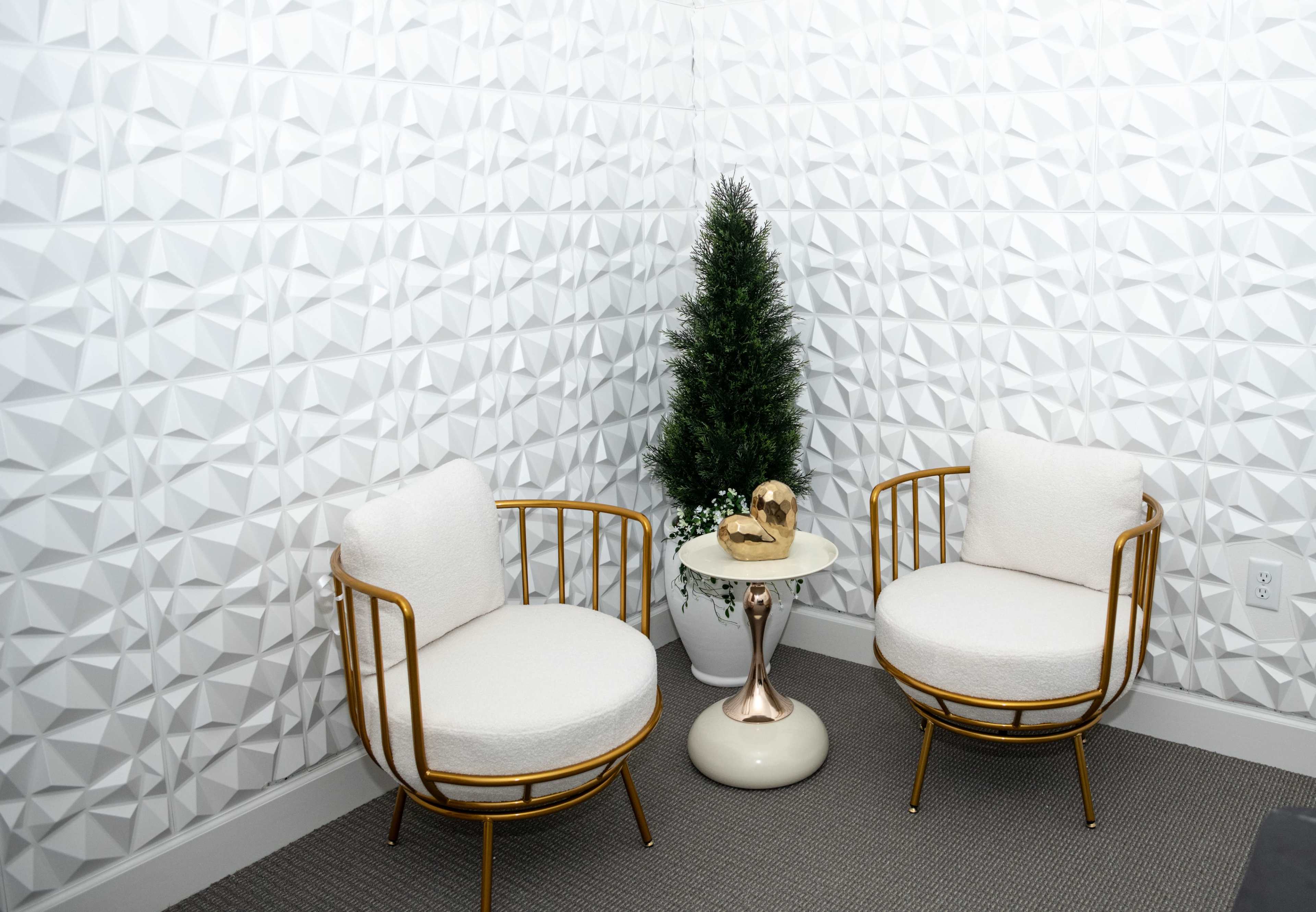 The scene features two upholstered chairs with gold frames positioned near a small side table and a potted artificial tree in a corner with textured, white walls.