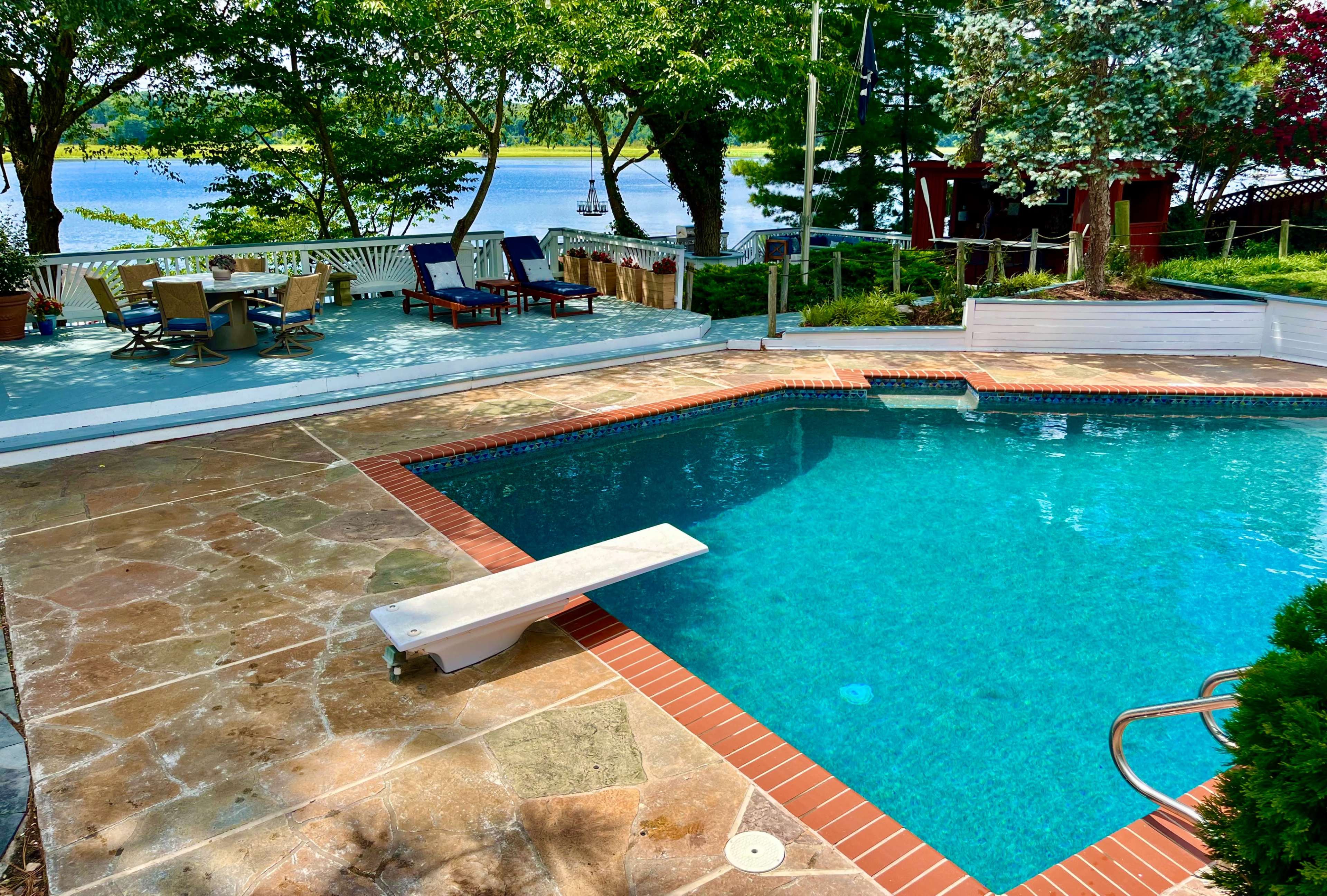 A swimming pool with a diving board is surrounded by a patio area that includes lounge chairs and overlooks a lake.