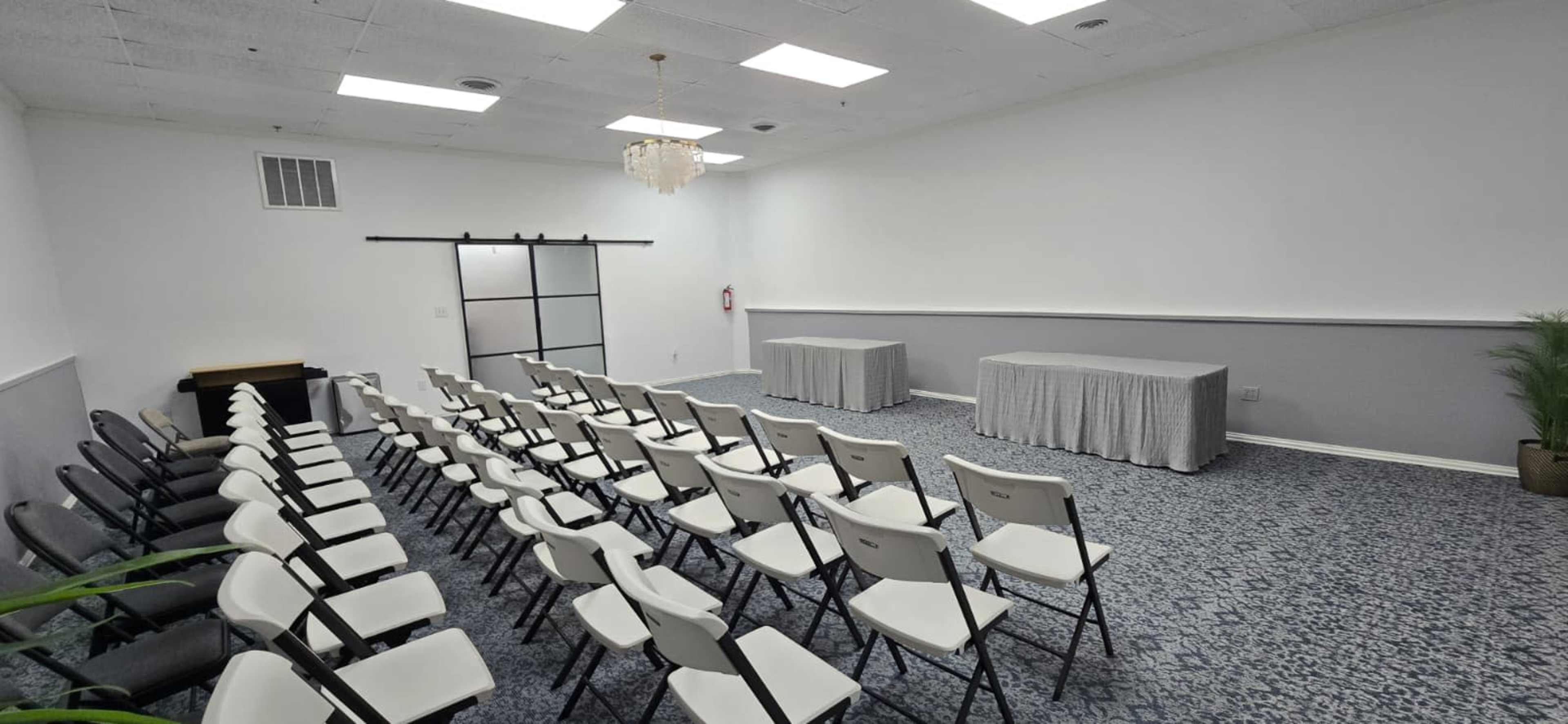 The image shows a small, empty conference room arranged with rows of chairs facing two tabled fronts.