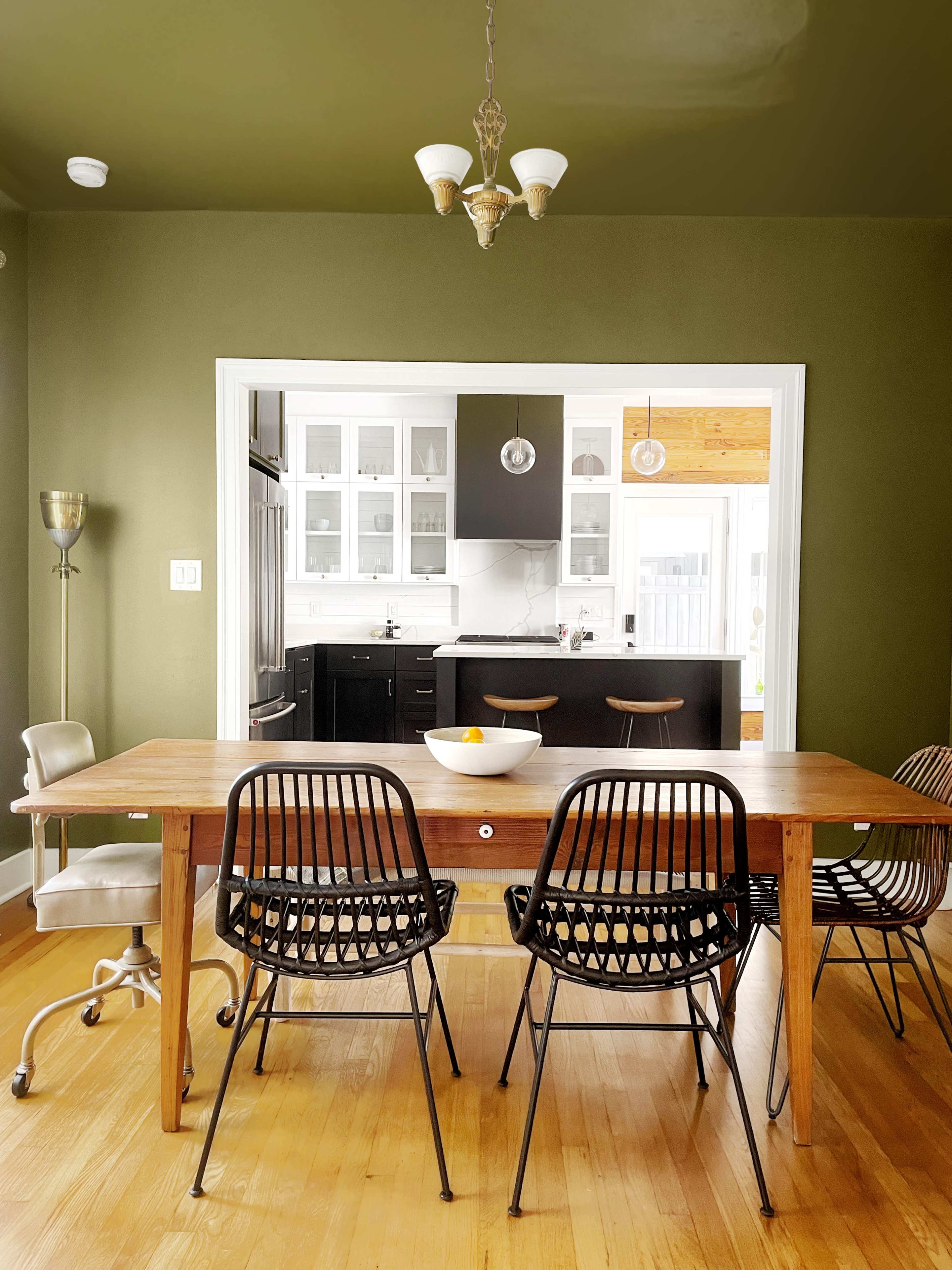 A wooden dining table surrounded by black wire chairs is positioned in a room with olive green walls, leading to a modern kitchen in the background.
