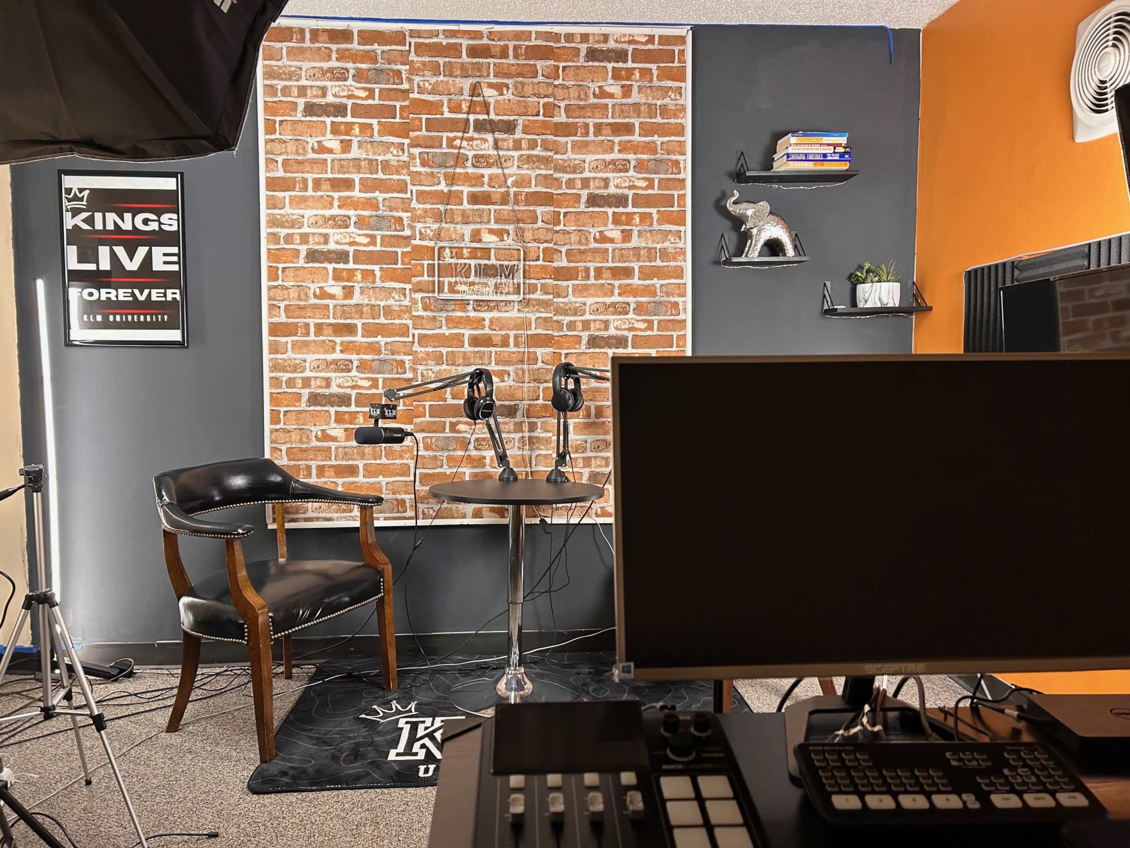 A podcast or interview setup featuring a black chair, a round table with two microphones, and a brick wall backdrop, with desert orange and dark gray sections around.