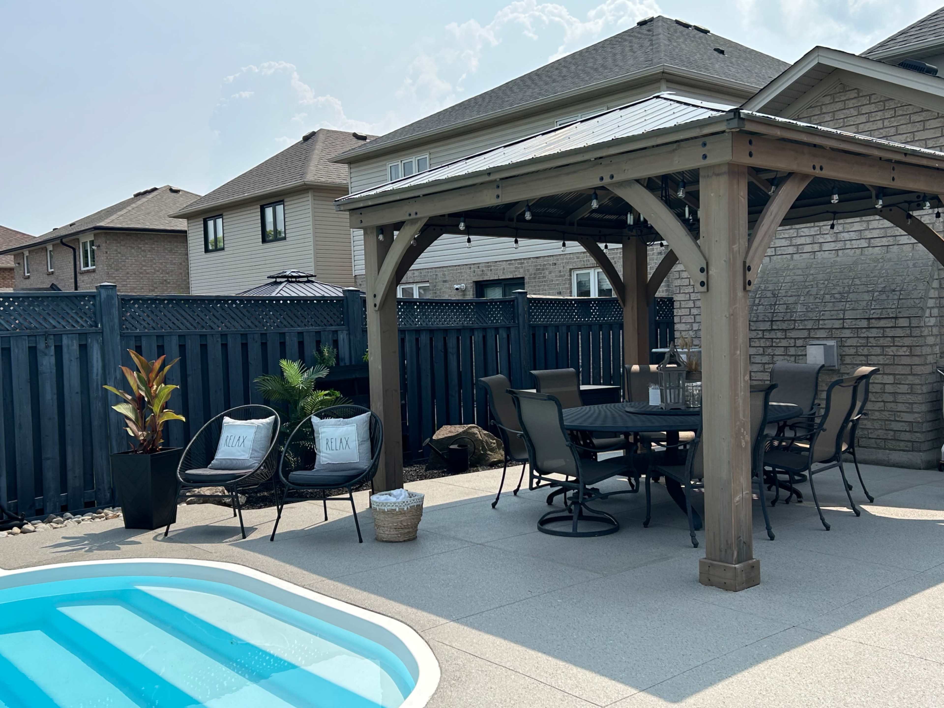 A backyard patio area featuring a wooden gazebo, a dining table, and a small swimming pool.